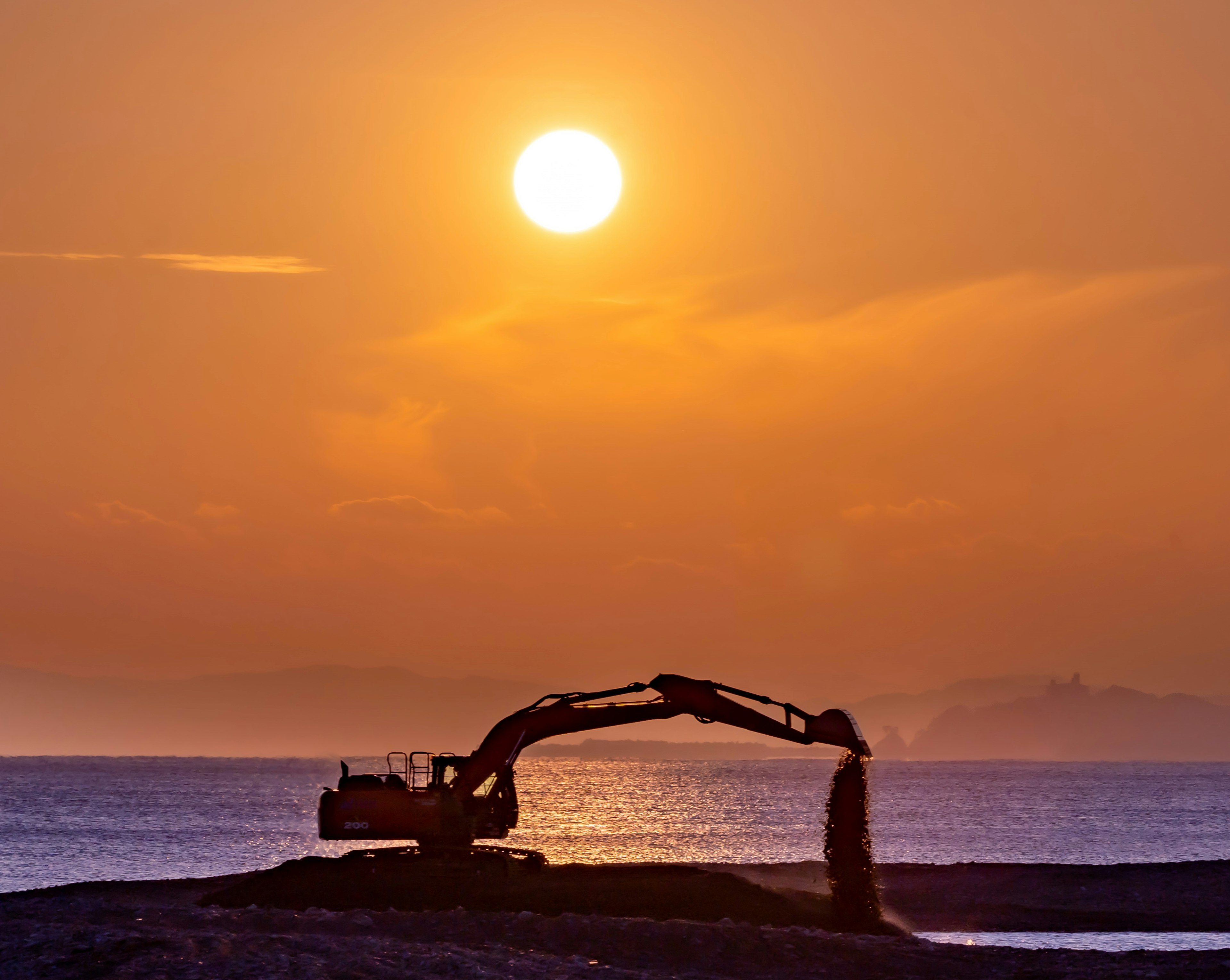 夕日を背景にした海岸で作業する掘削機