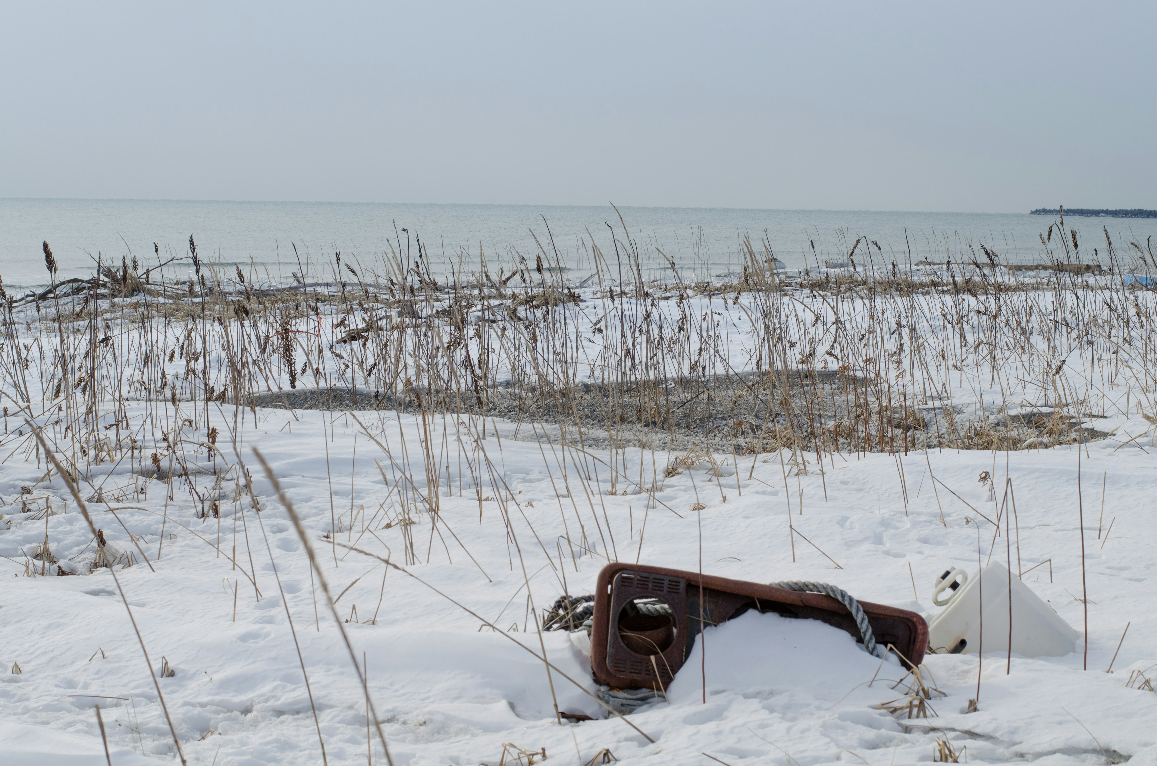 Verlassener alter Stuhl an einem verschneiten Strand mit spärlichem Gras und ruhigem Wasser