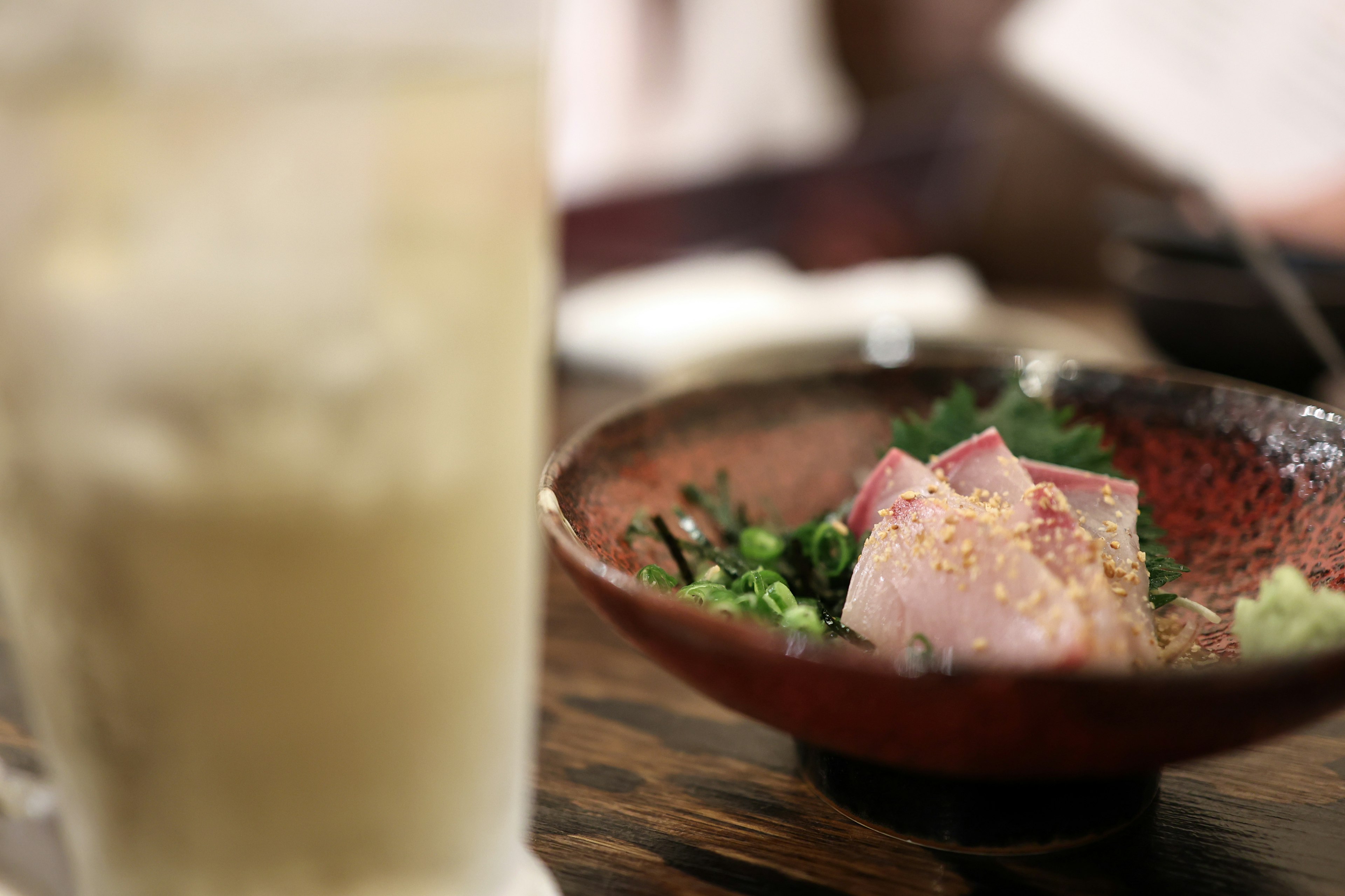 A plate of sashimi and greens on a table with a drink in the background