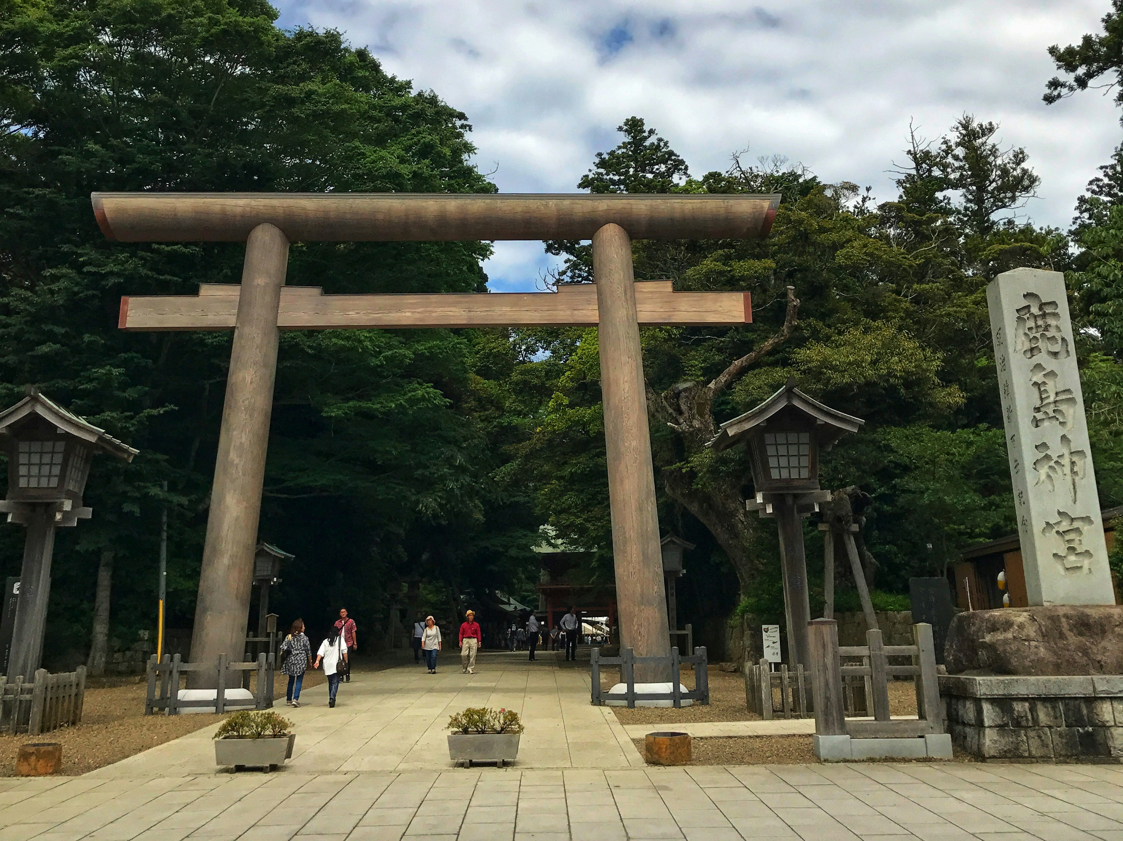 Một cổng torii lớn và bia đá tại lối vào đền với khách tham quan đang đi bộ