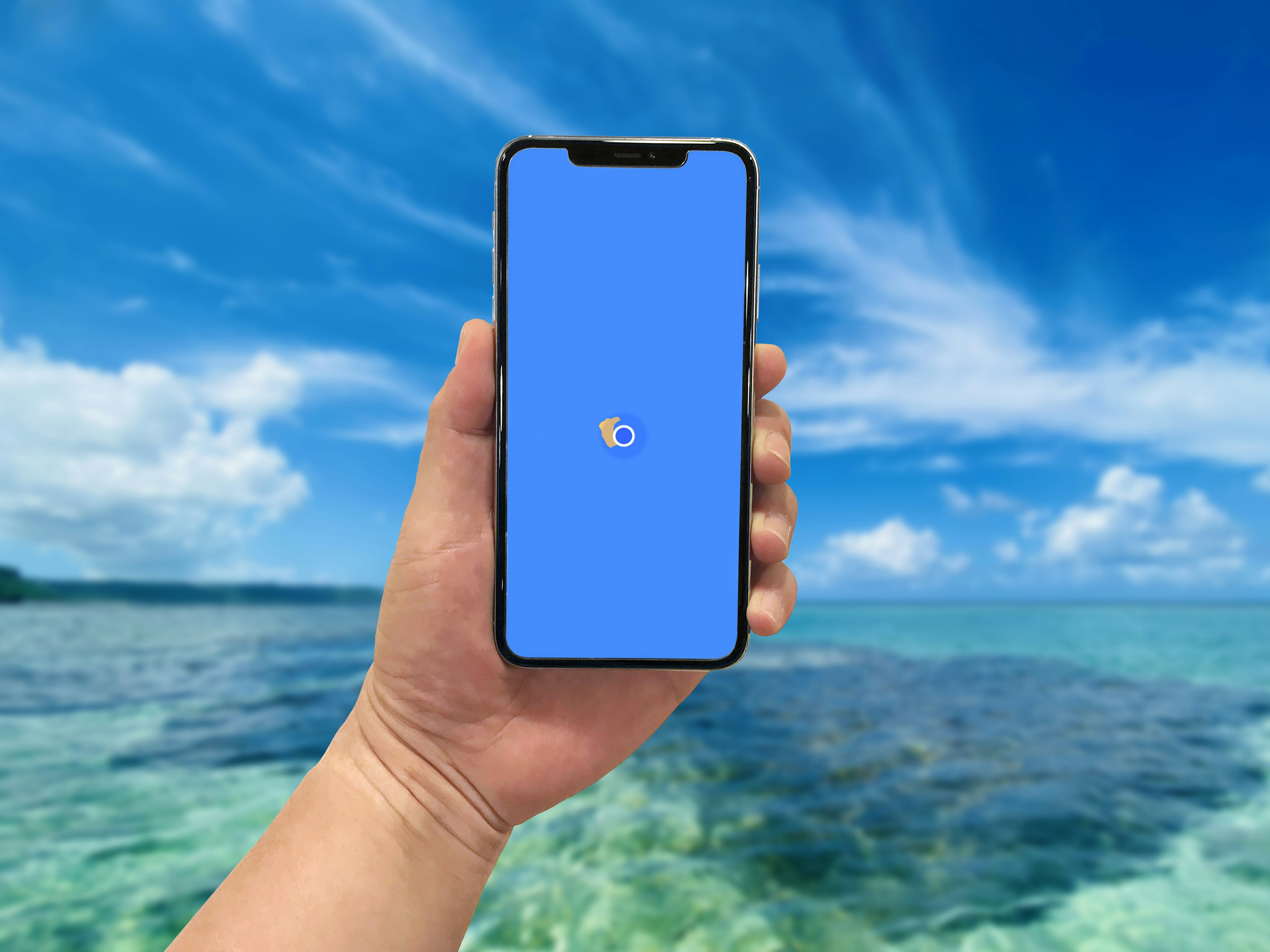 Sebuah tangan memegang smartphone dengan layar biru di depan lautan yang jernih