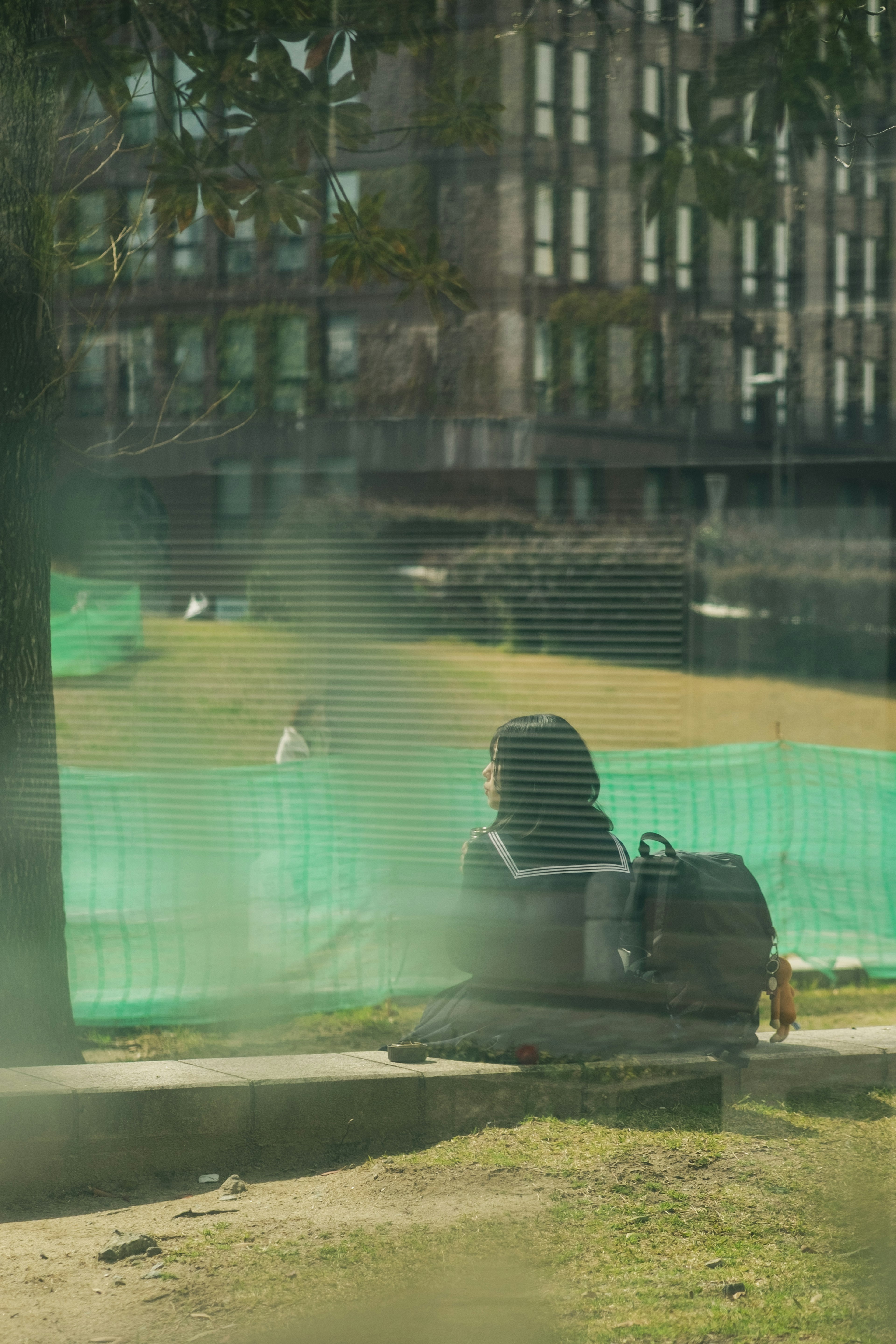 A person sitting near a green net in a park with buildings in the background