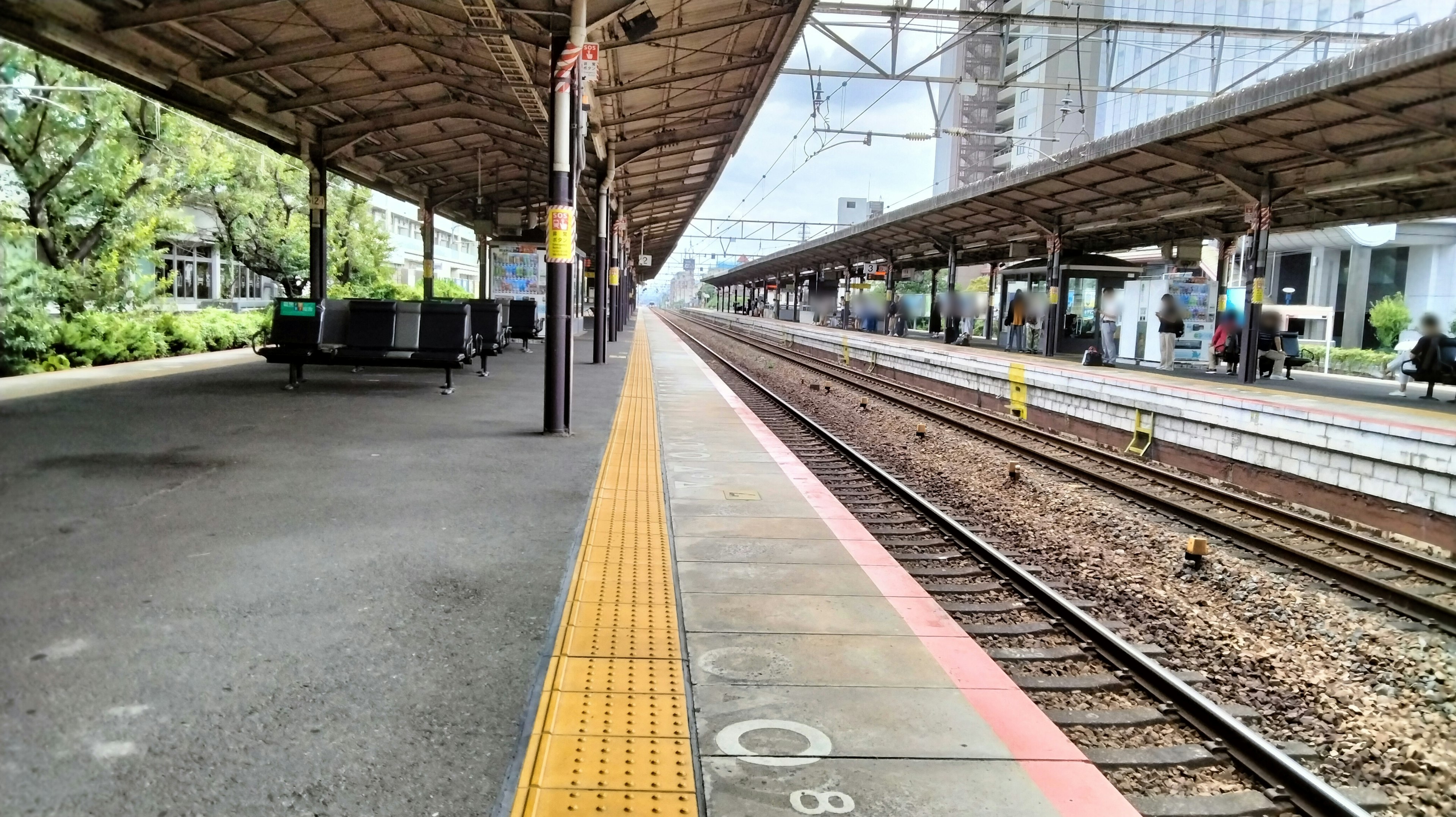 View of a train station platform and railway tracks featuring the station structure and surrounding greenery