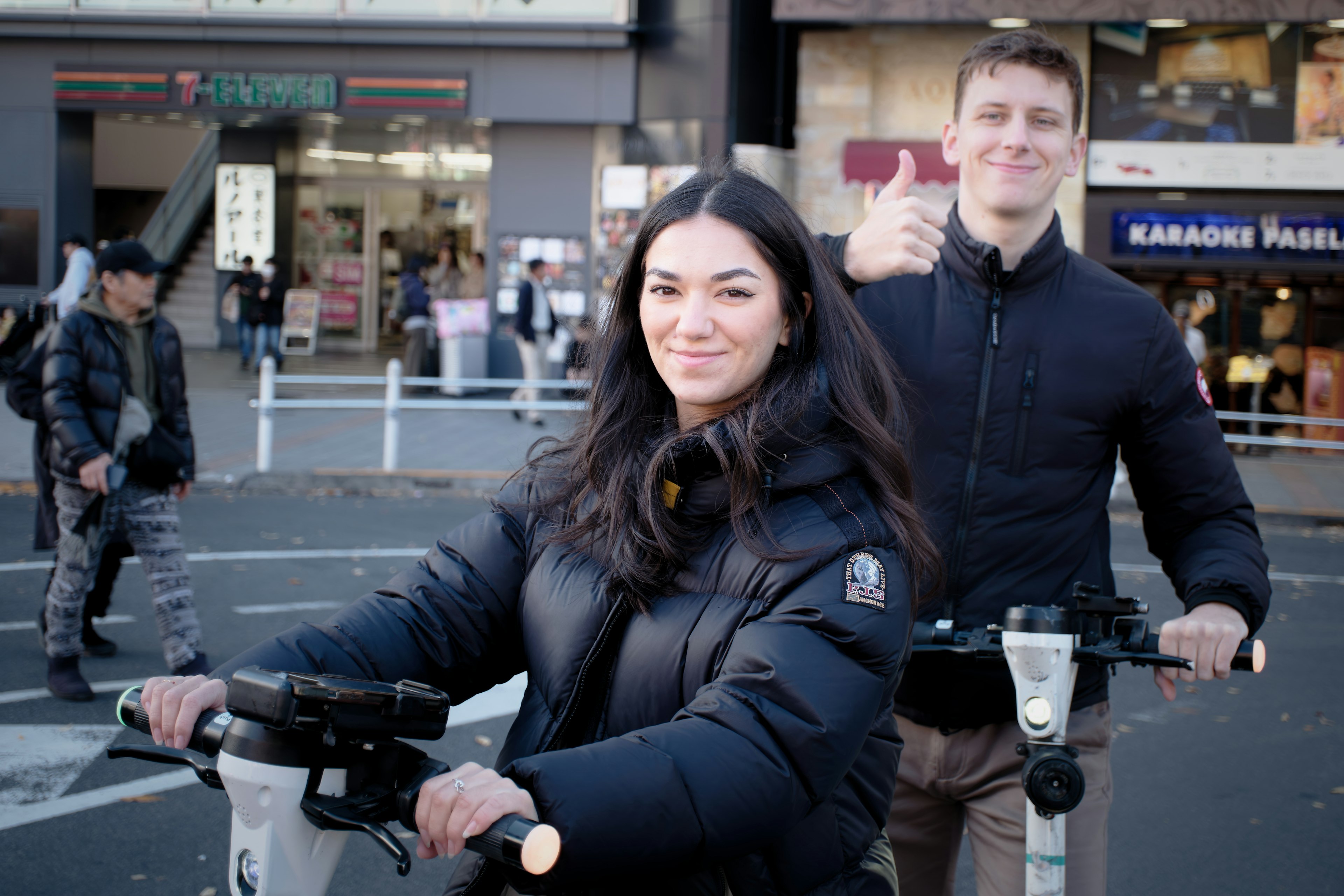 Dos jóvenes montando scooters eléctricos, mujer con chaqueta de plumas negra, hombre sonriendo y haciendo un pulgar arriba