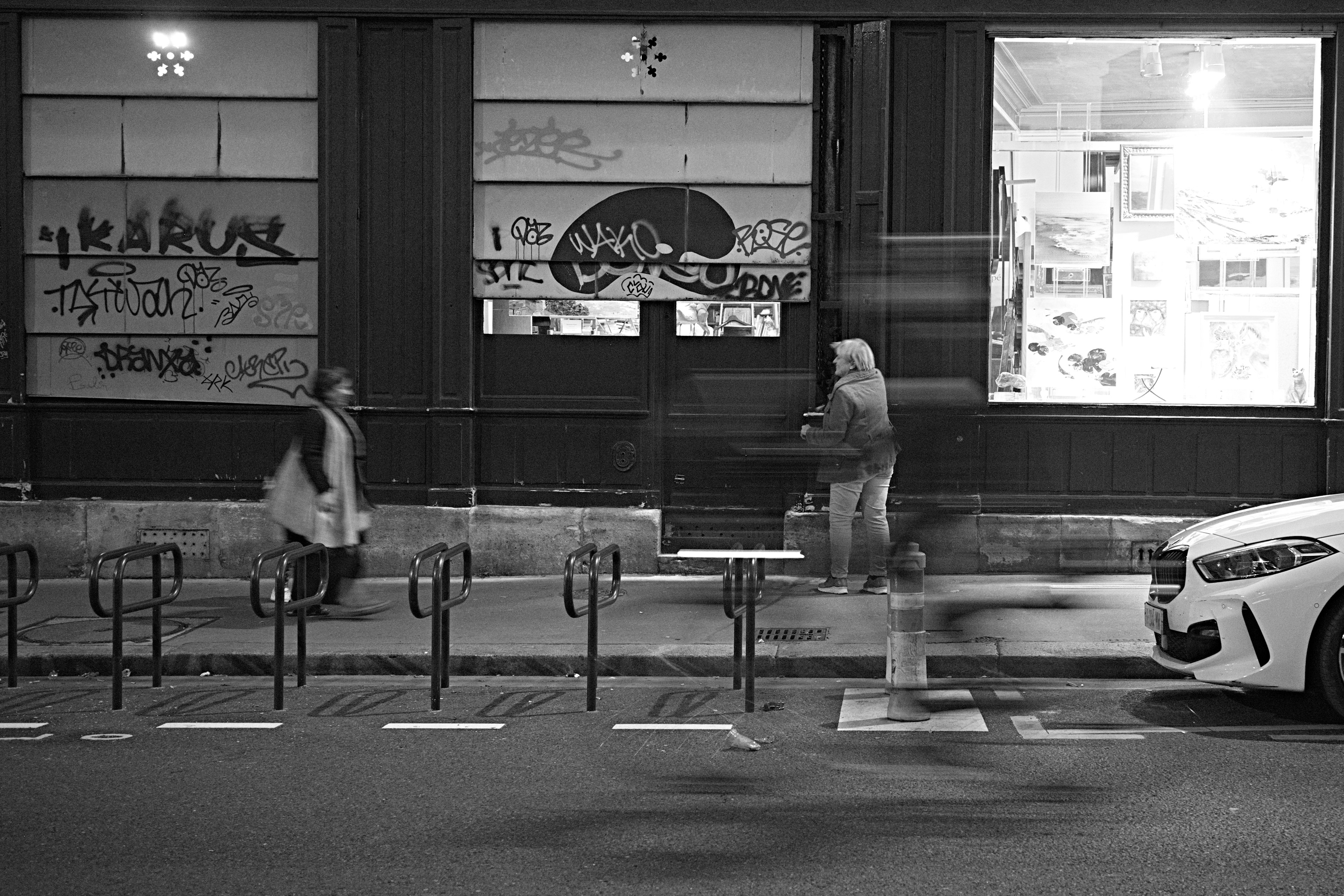 Black and white street scene with pedestrians and cyclists passing by Graffiti on walls and bright store interior visible through windows