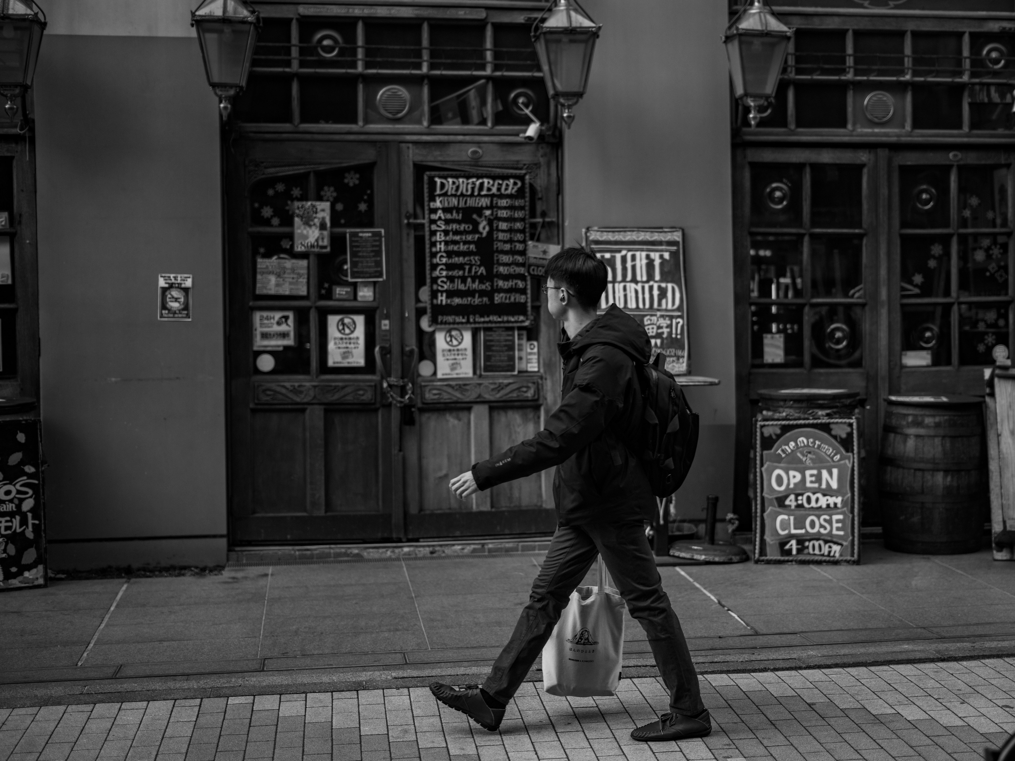 Un hombre caminando en una escena de calle en blanco y negro frente a una tienda con un cartel de abierto