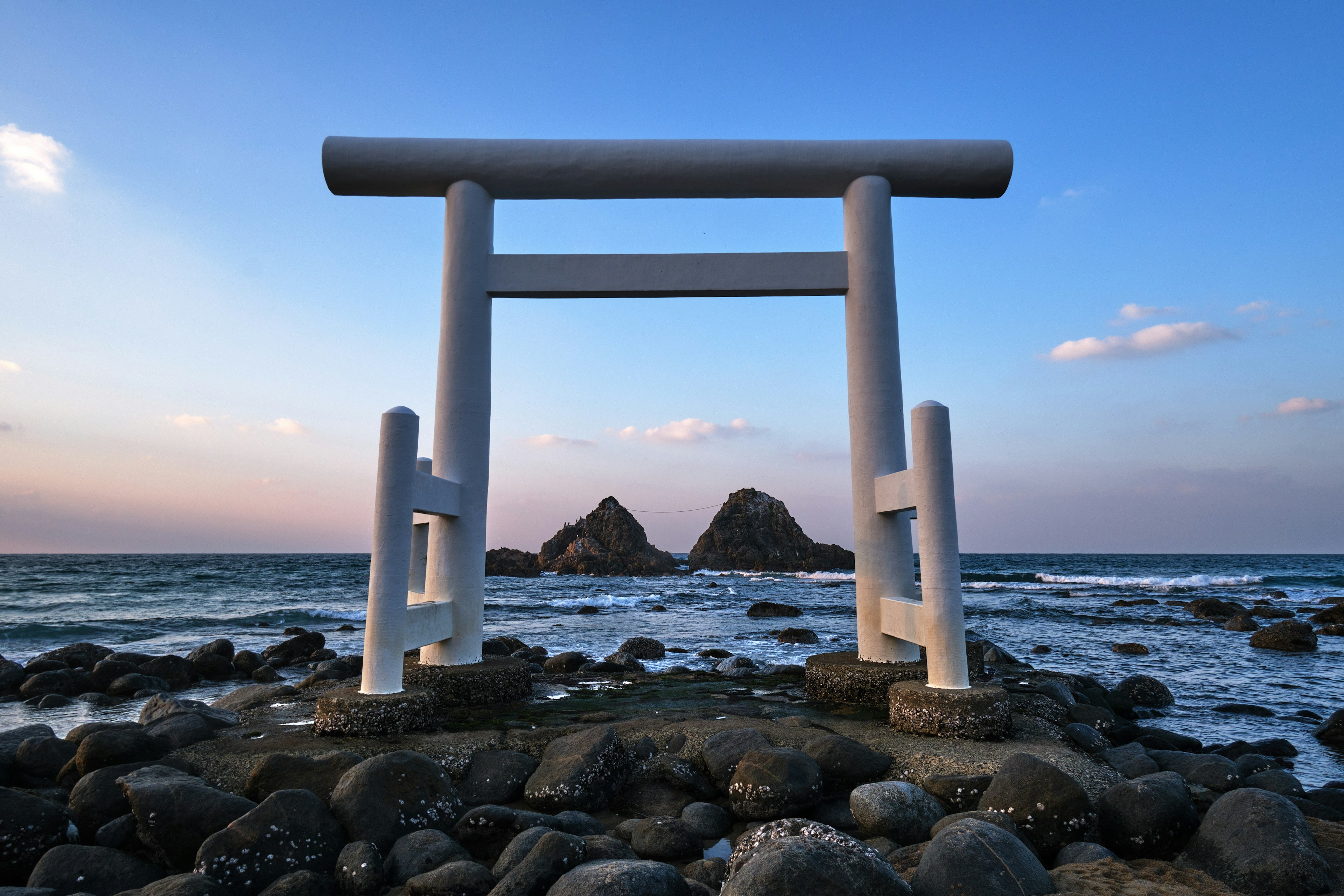 Pintu torii putih dengan pemandangan laut saat matahari terbenam menciptakan suasana tenang