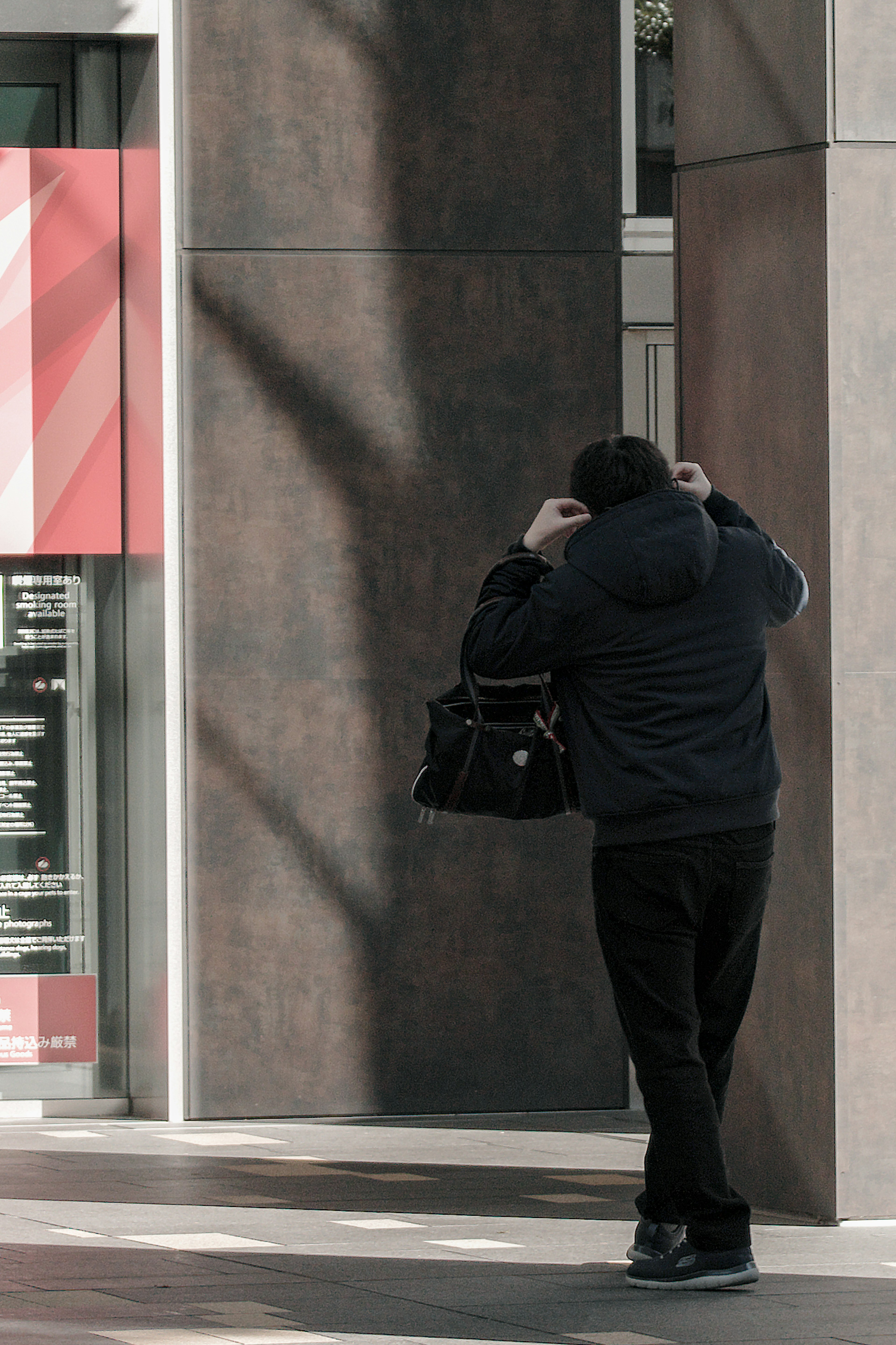 Persona in piedi di spalle contro un muro con abbigliamento scuro che tiene una borsa ombre sottili in un contesto urbano