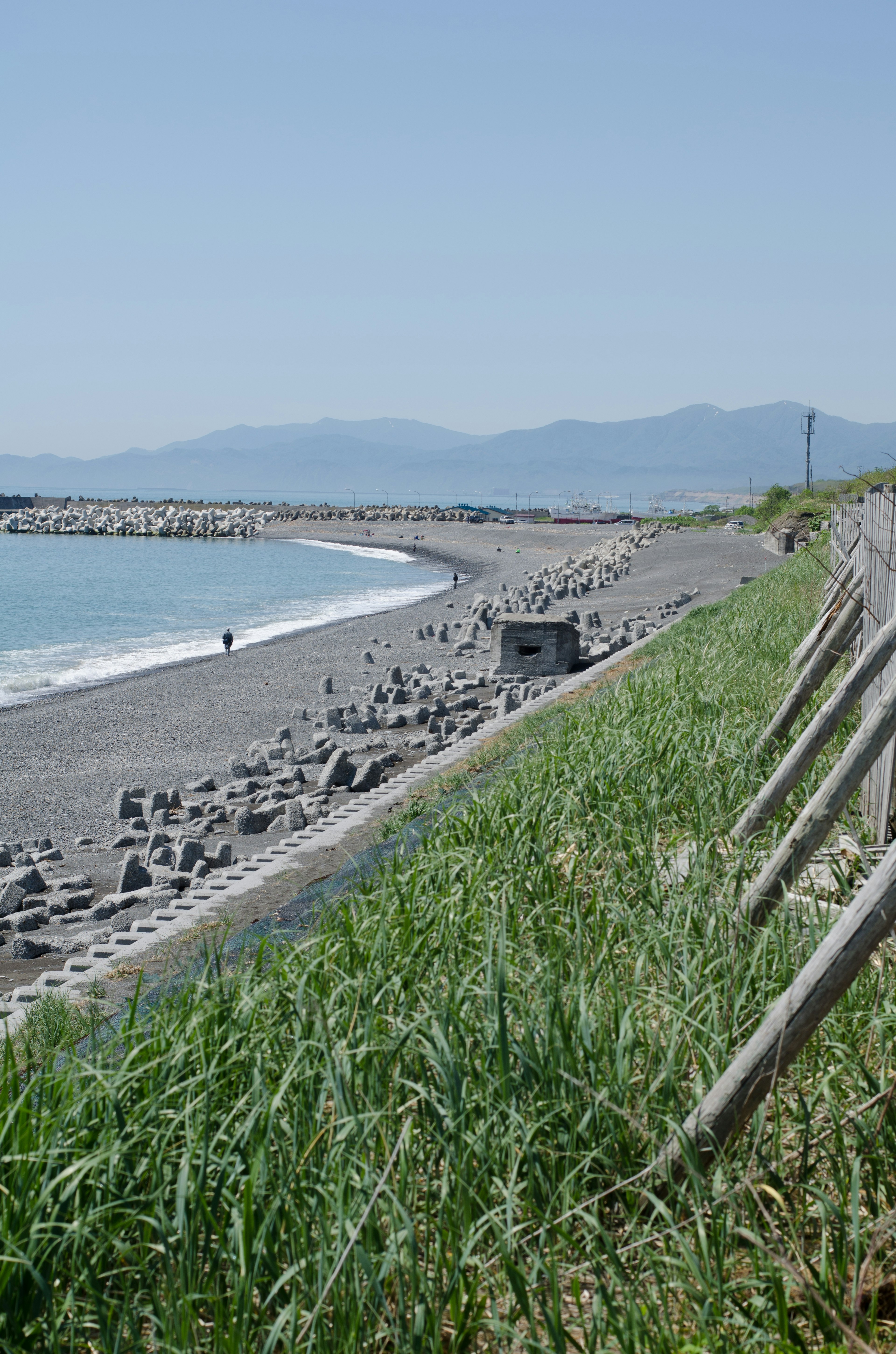 Strandansicht mit blauem Ozean und Bergen im Hintergrund mit grünem Gras und Kies