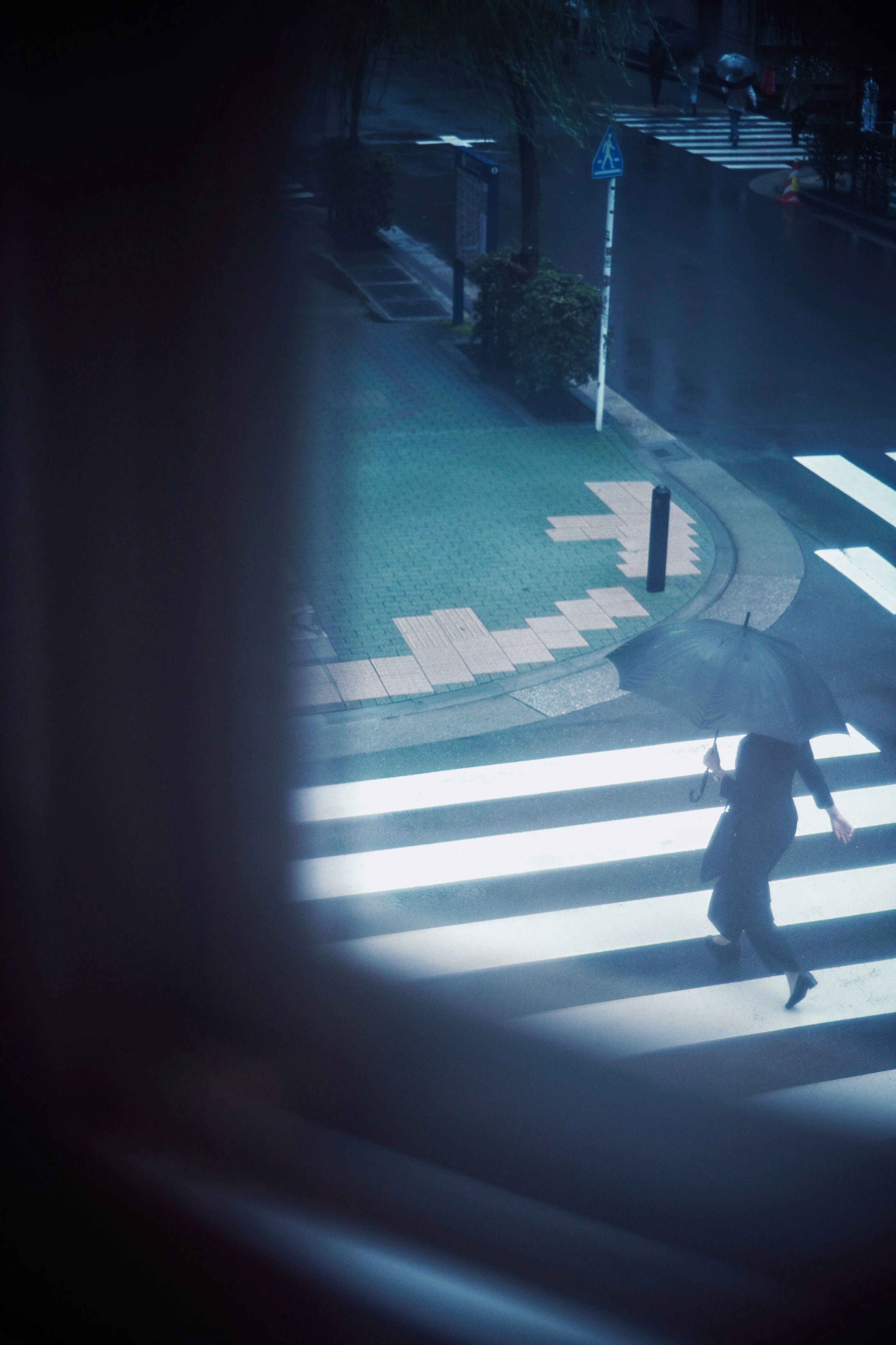 青いトーンの街で傘をさした人物が横断歩道を渡っている