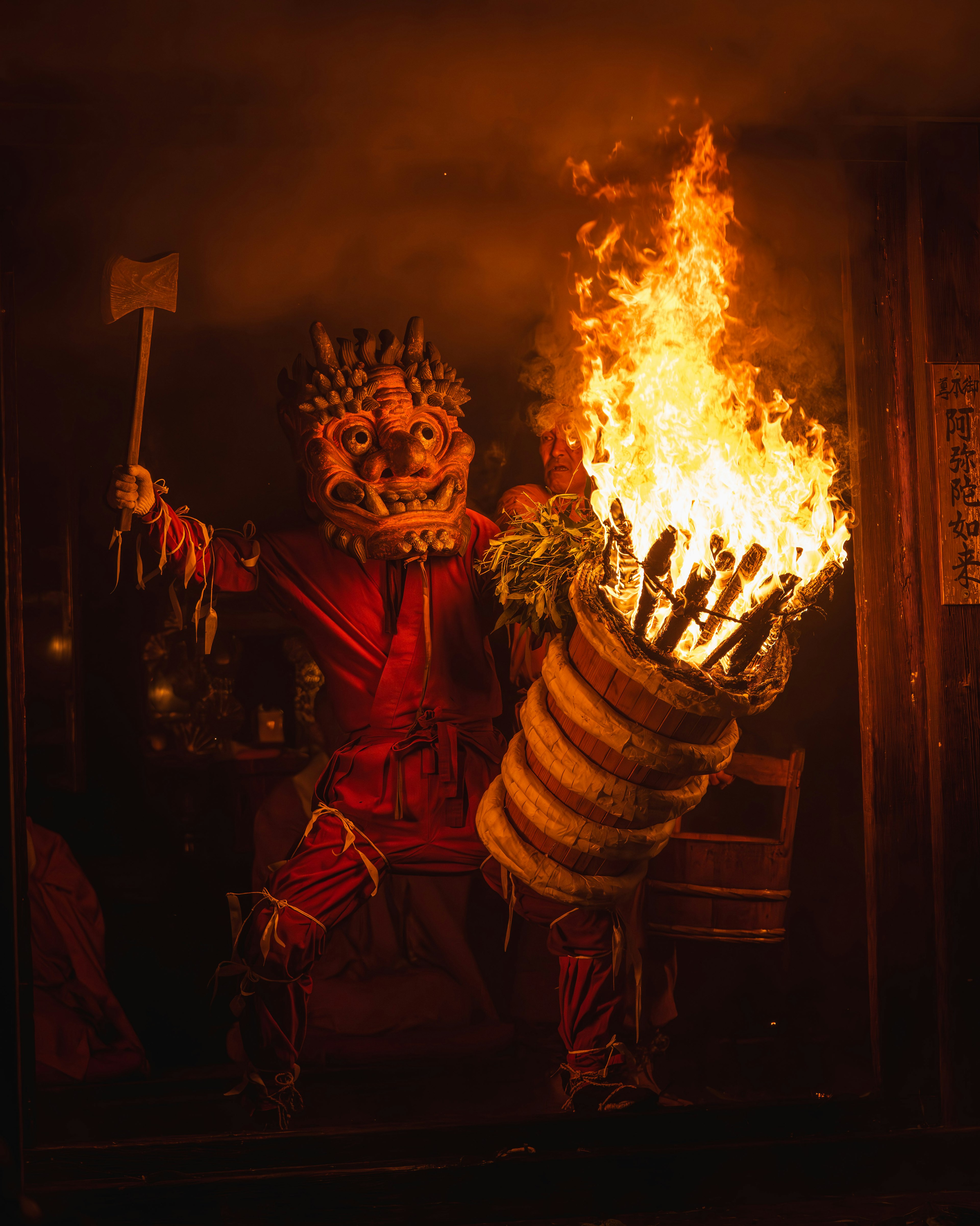 Eine maskierte Figur in roter Kleidung, die eine brennende Fackel und einen Holzbehälter hält