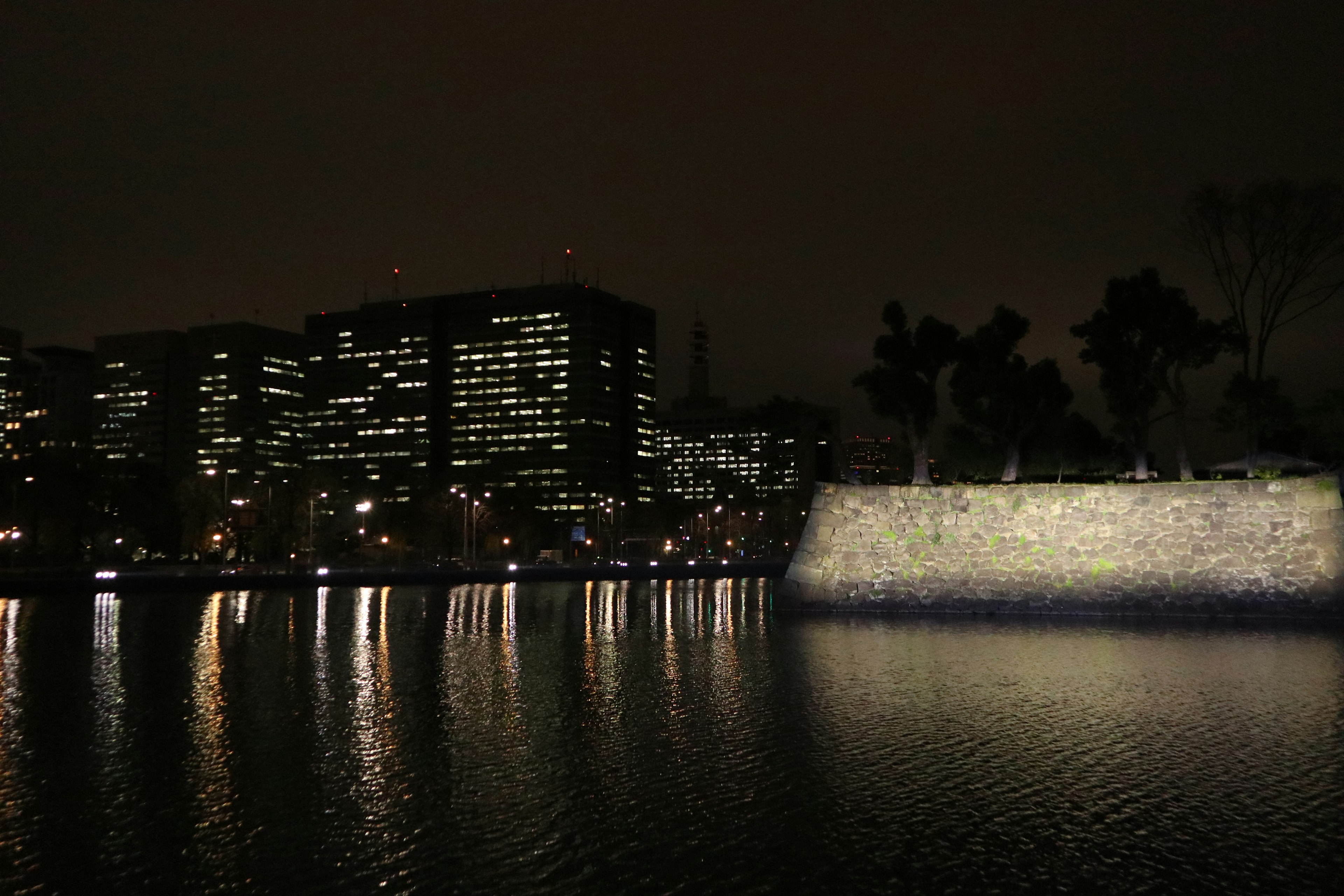 Paisaje urbano nocturno con reflejos de edificios en el agua y un muro de piedra iluminado