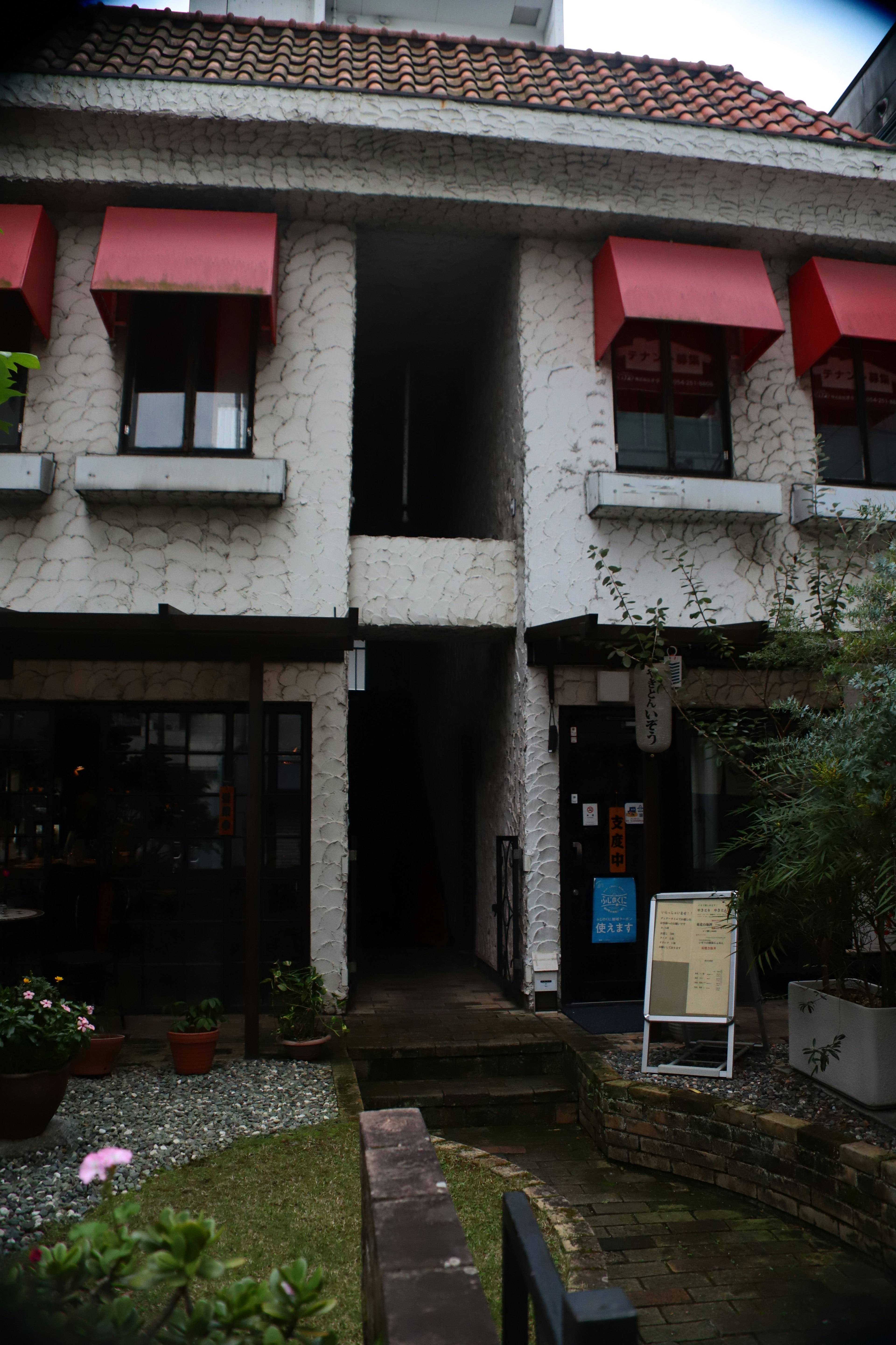 Entrance of a white building with red awning windows surrounded by plants