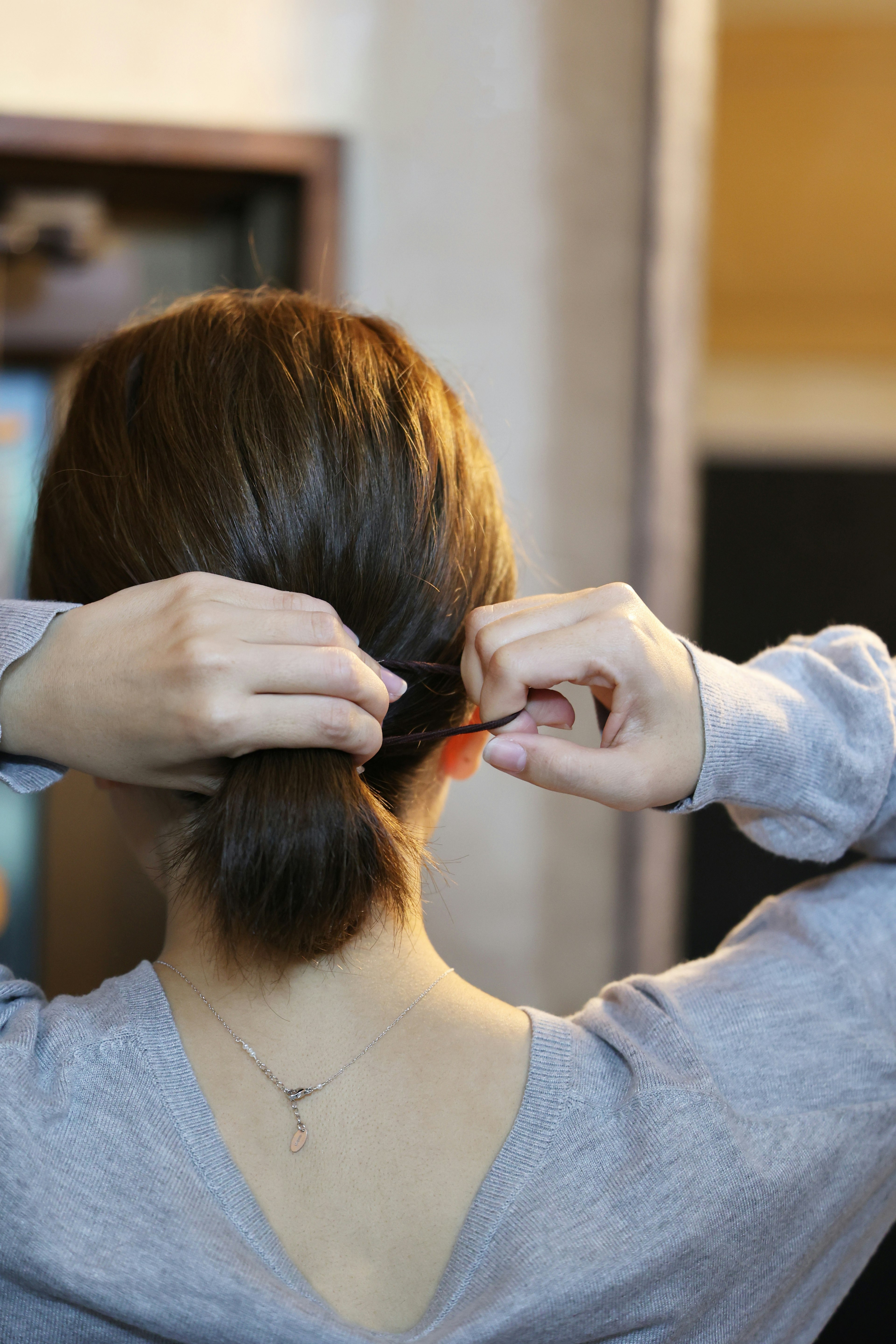 Woman tying her hair back seen from behind wearing a gray long-sleeve shirt using a hair accessory