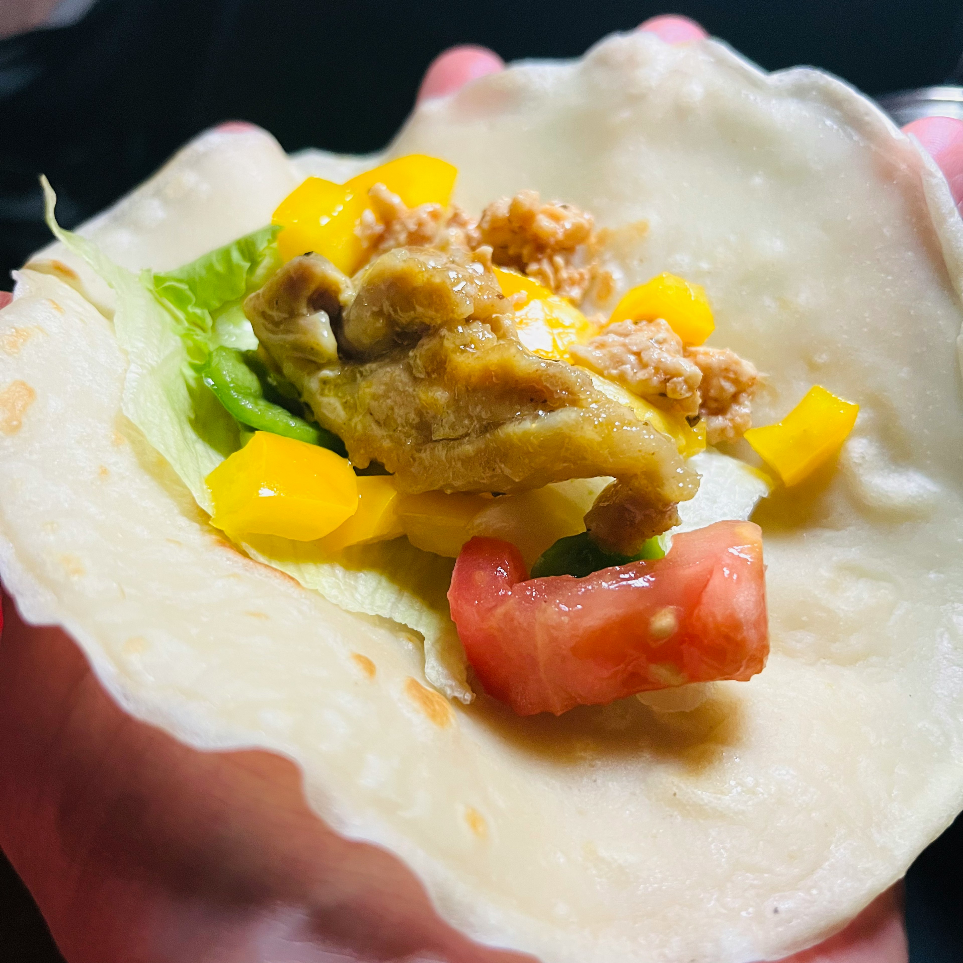 Un taco tenu par la main contenant de la laitue, des tomates, des poivrons jaunes et du poulet