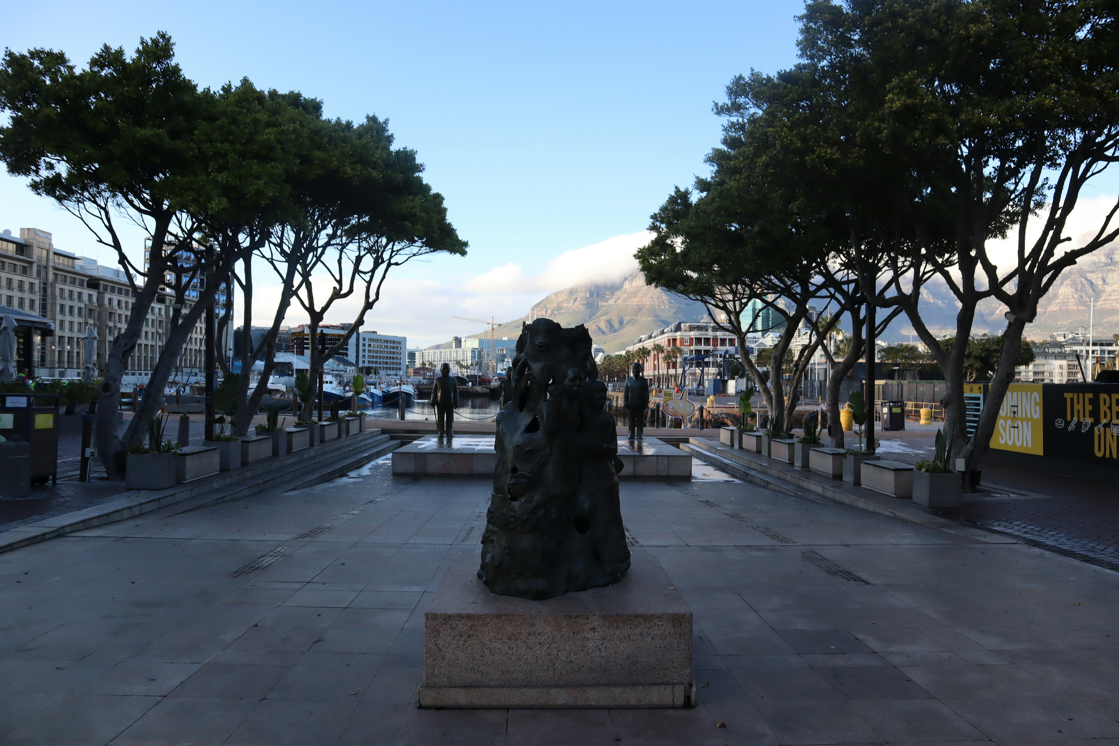 Escultura de piedra en una plaza rodeada de árboles verdes con montañas al fondo