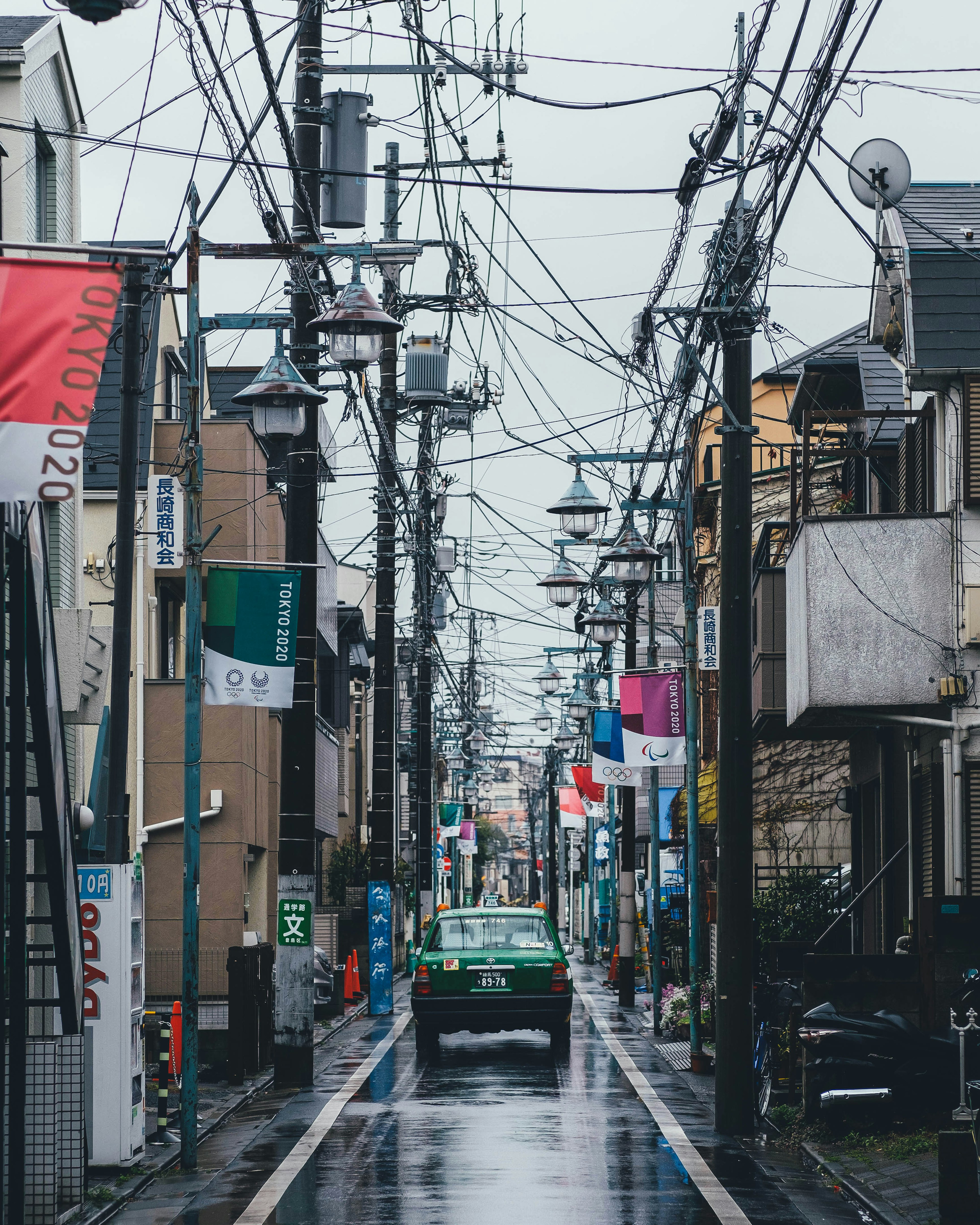 Vista di una strada con un'auto verde in una giornata di pioggia circondata da linee elettriche e bandiere colorate