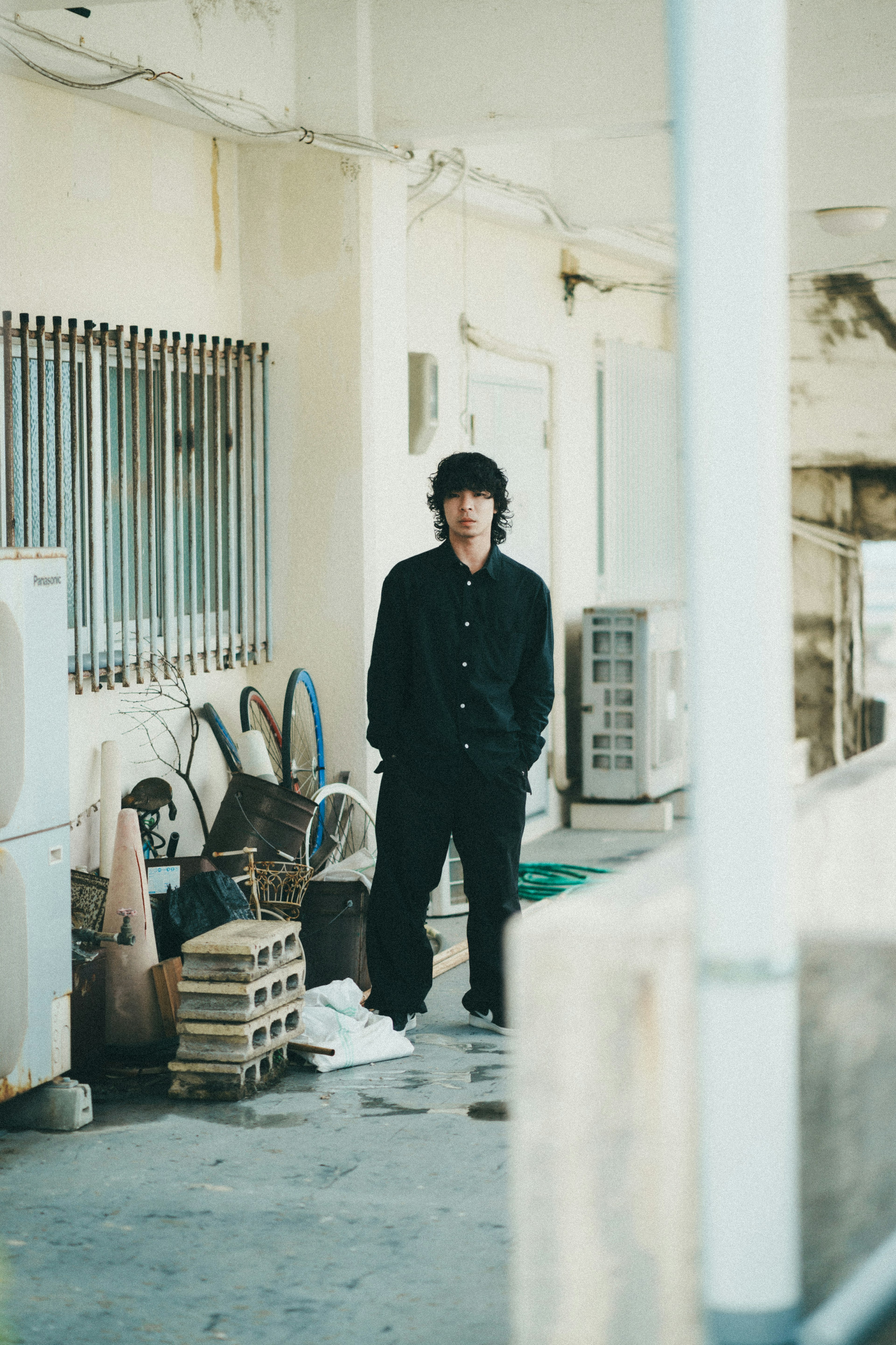 Young man standing in an outdoor corridor wearing black clothing surrounded by scattered objects