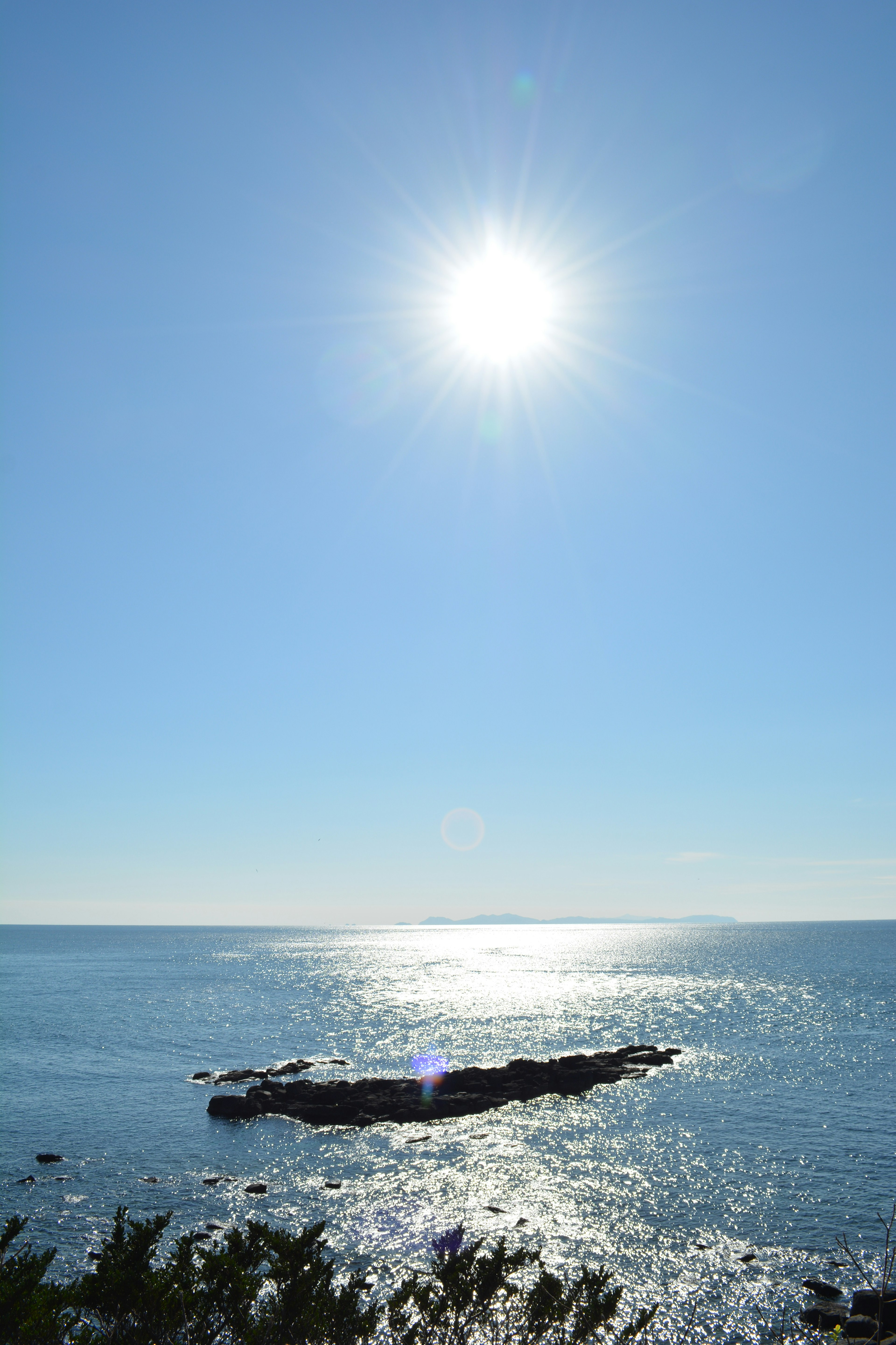 Bright sun over a serene blue ocean with a rocky shoreline