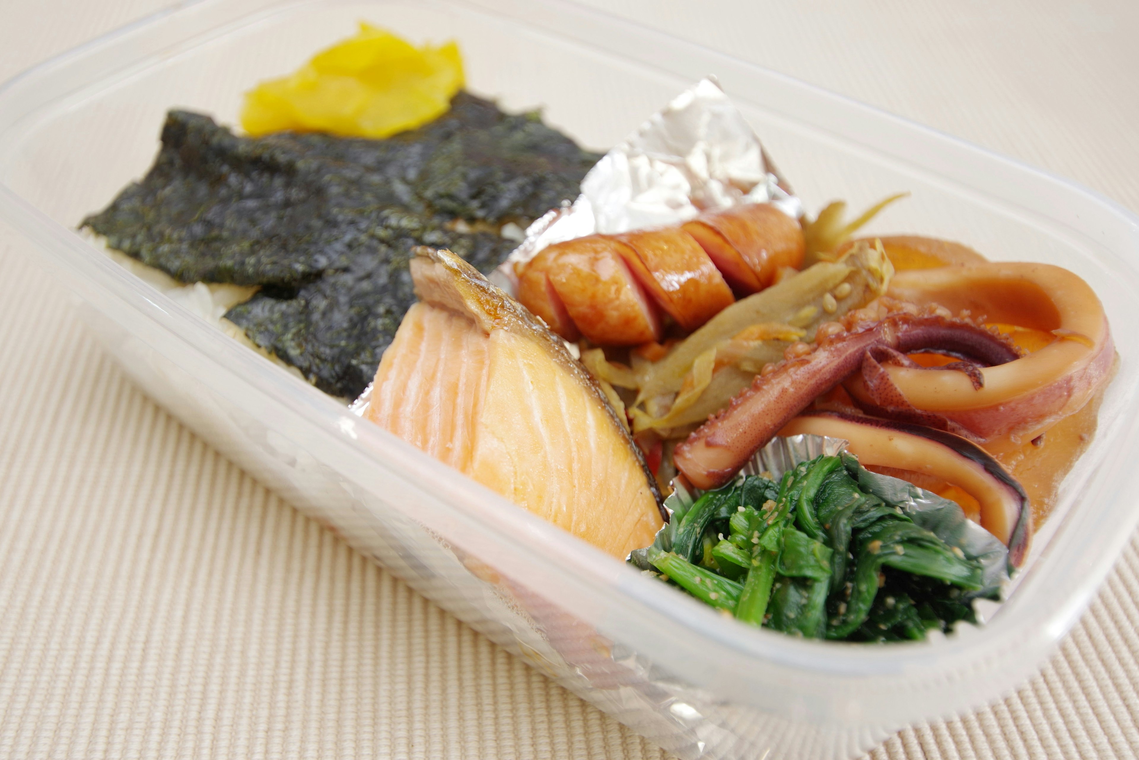 Japanese bento box featuring salmon, seaweed, vegetables, squid, and sausage