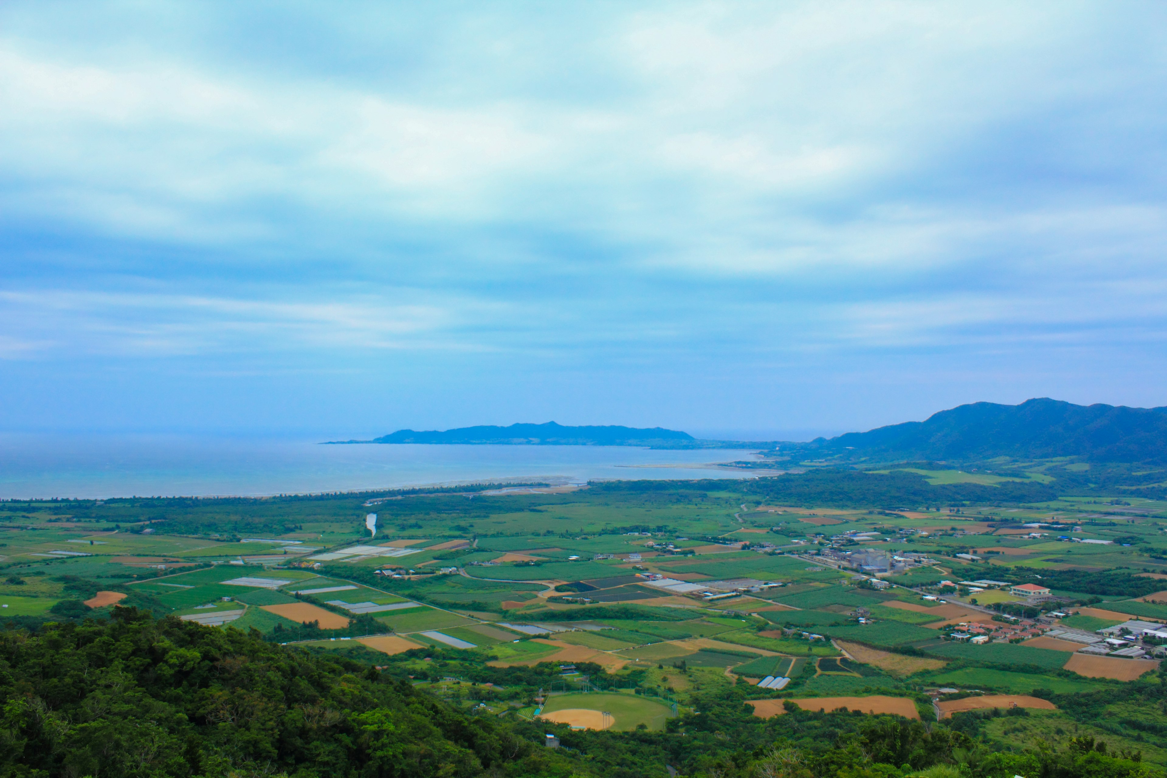 Vast green farmland under a blue sky with clouds and a view of the sea