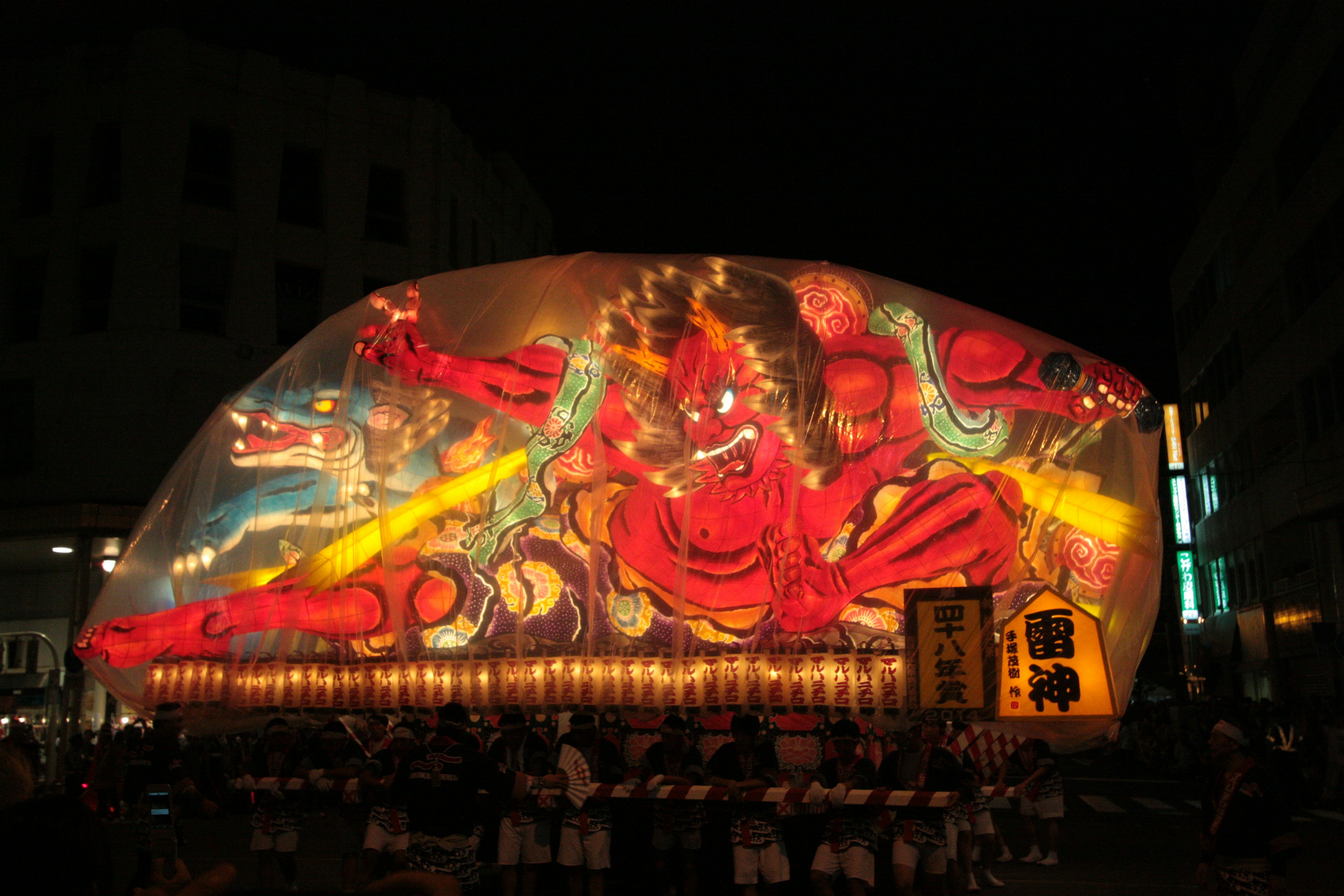 Eine große beleuchtete Laterne eines roten Dämons bei einem Nachtfestival mit umstehenden Menschen