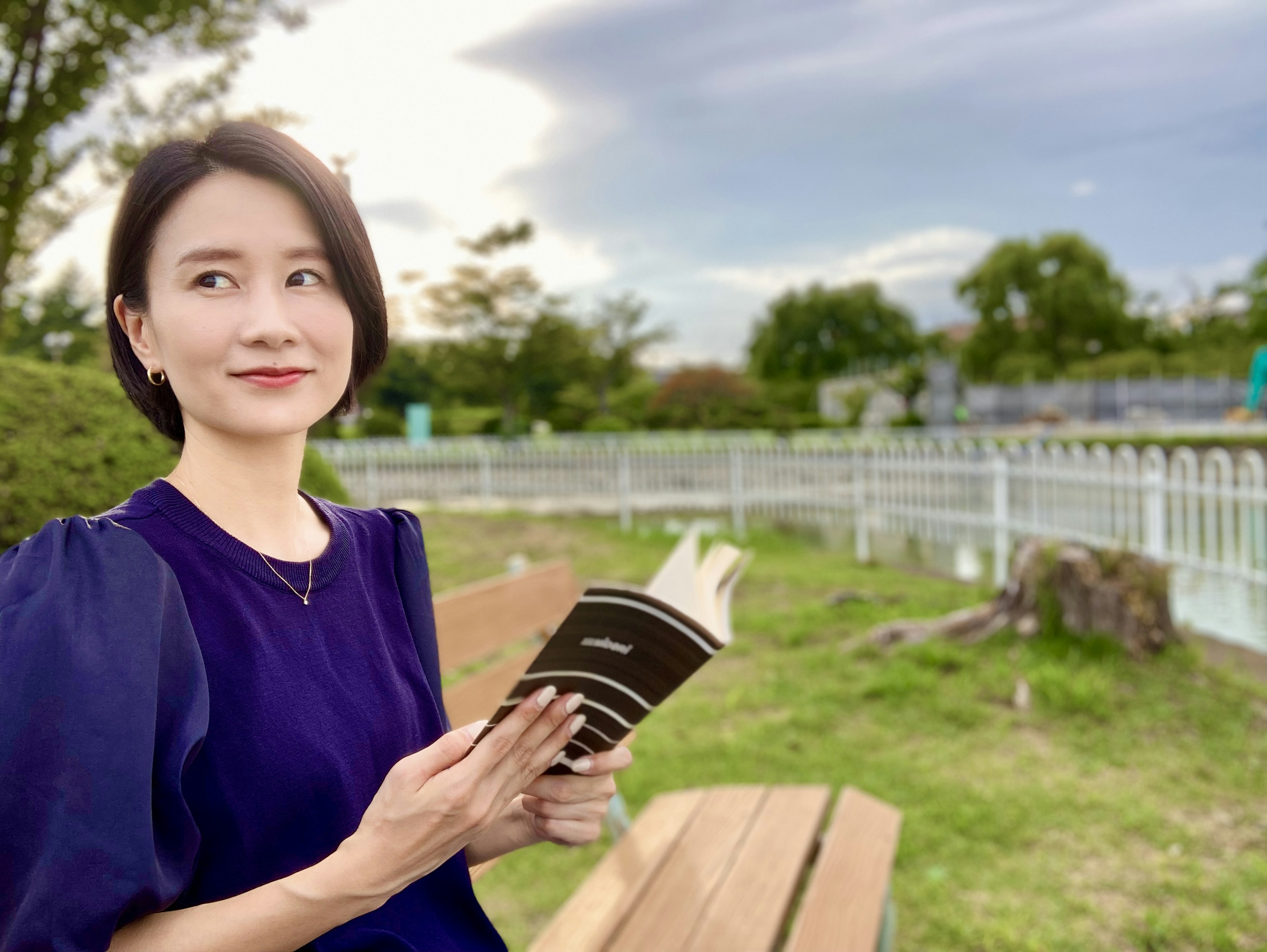 公園のベンチに座り、黒い本を持つ女性が微笑む風景