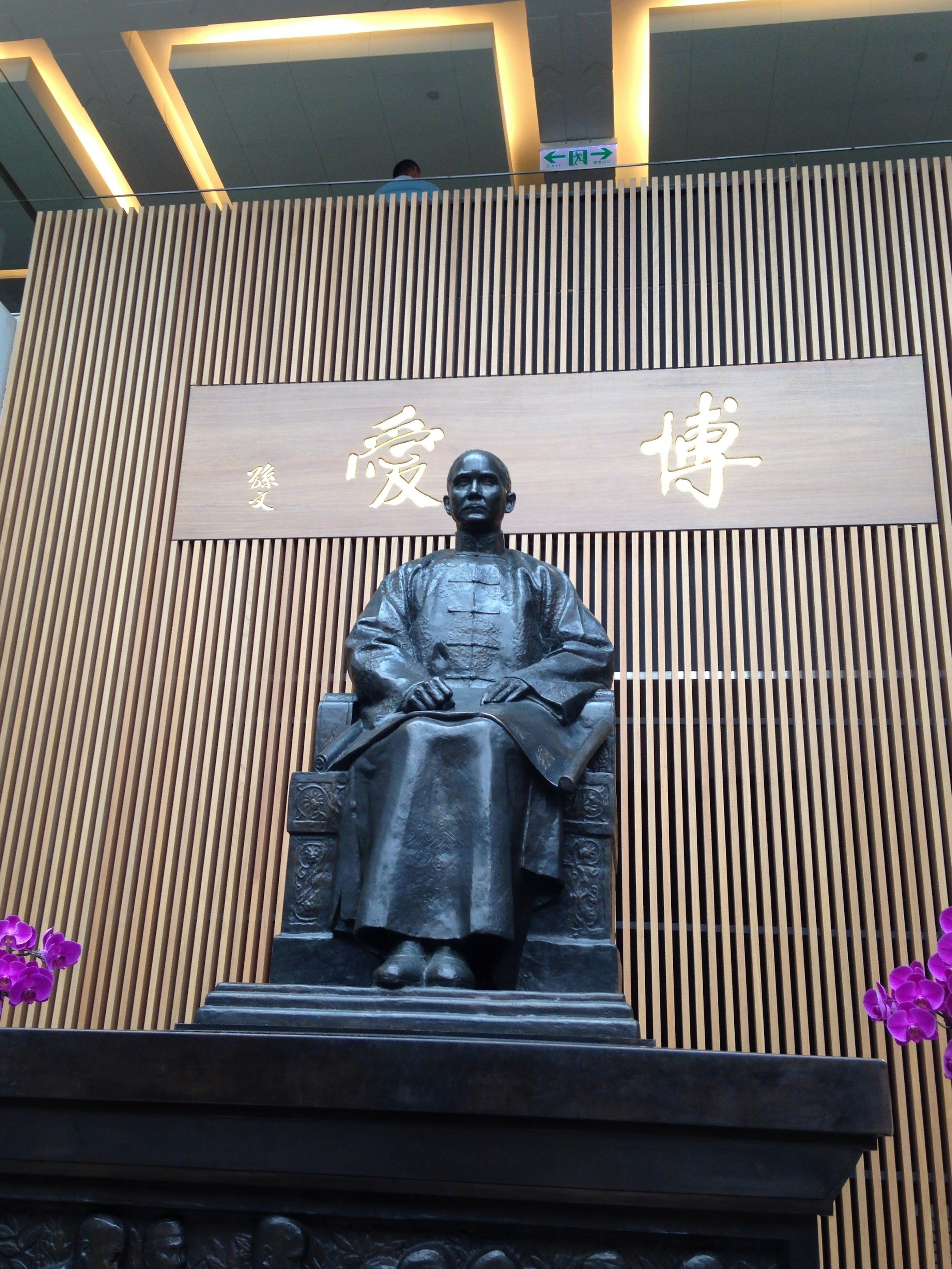 Estatua de bronce sentada con un muro de madera y flores moradas al fondo