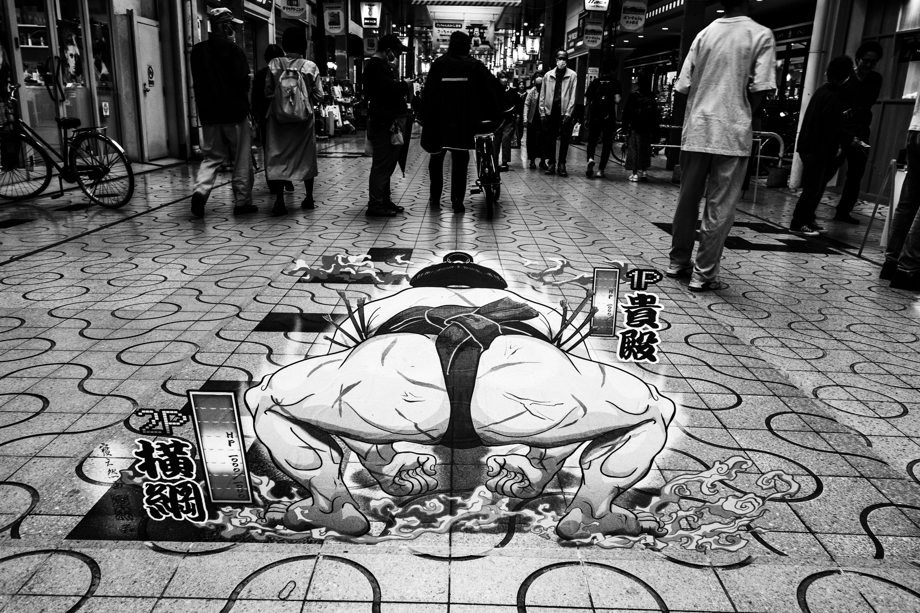 Black and white street art depicting a sumo wrestler lying down amidst pedestrians