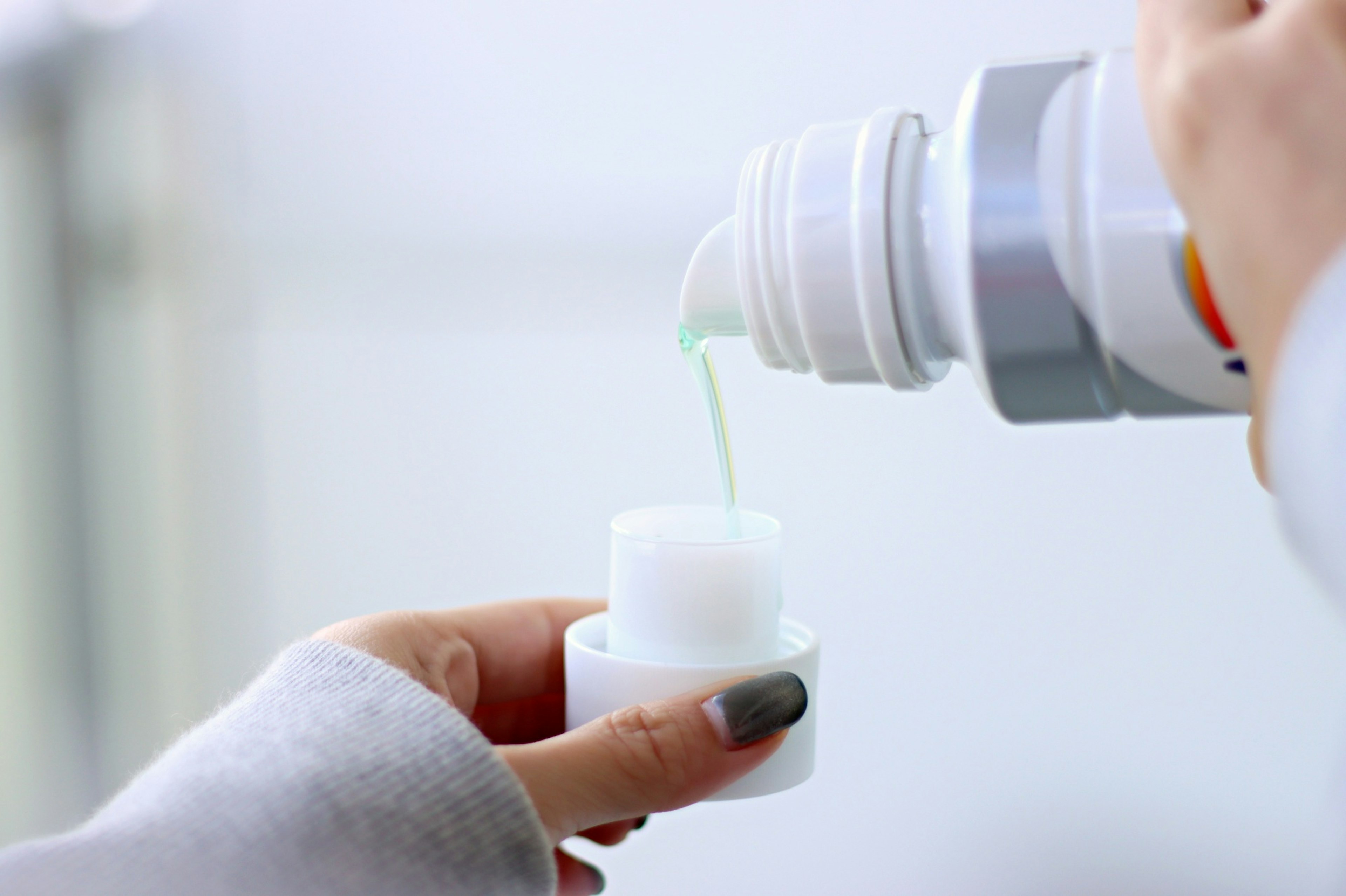 A hand pouring a transparent liquid from a white bottle into a small white cap with distinct fingernails