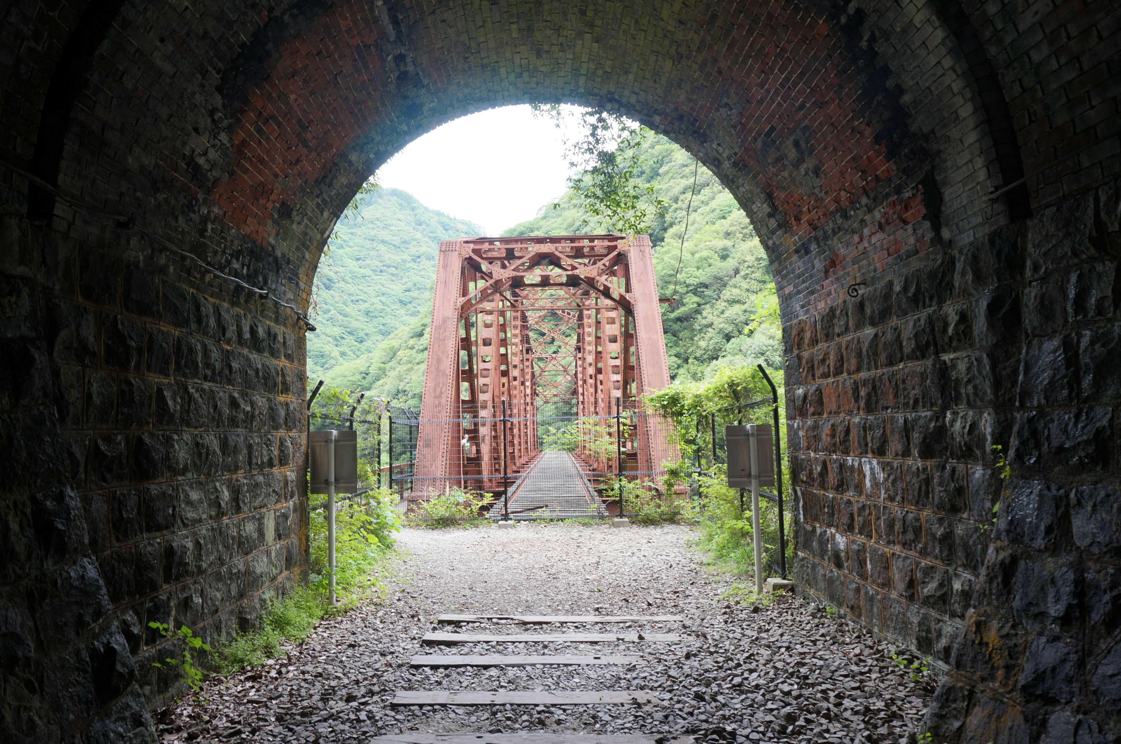 Blick auf eine rote Eisenbrücke durch einen Steintunnel mit umliegendem Grün