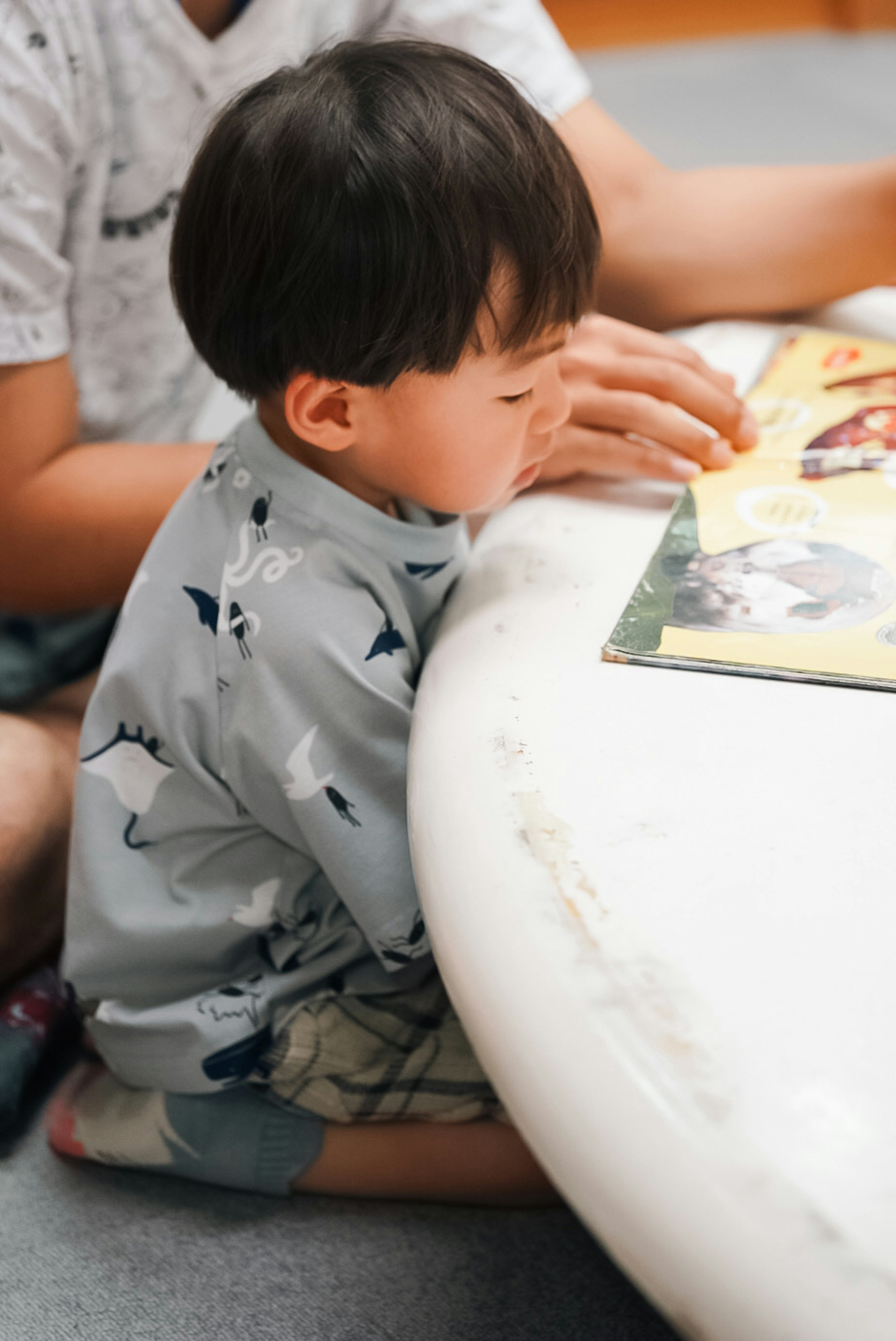 Seorang anak membaca buku sambil duduk di samping meja putih besar