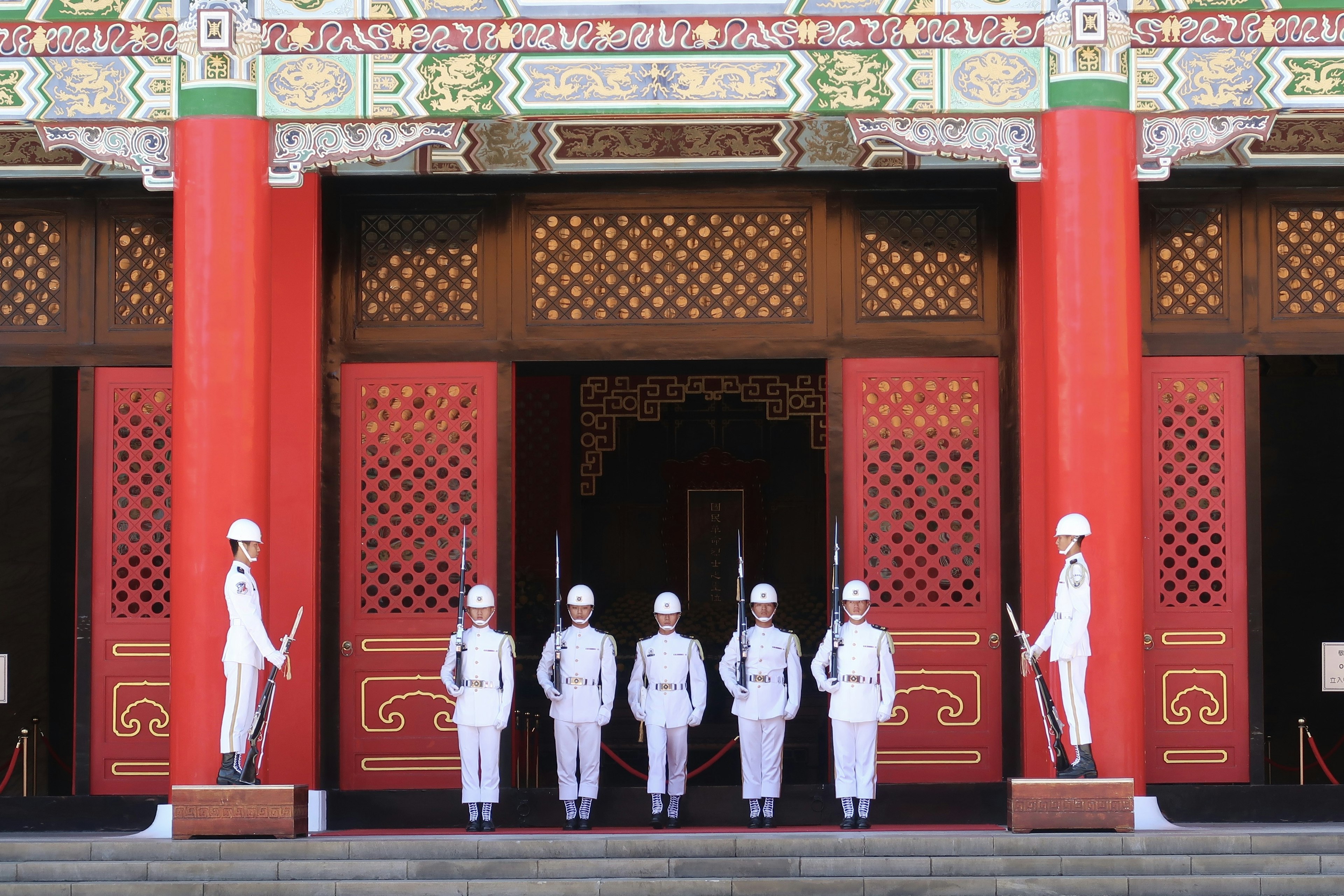 Guards in uniform standing in formation in front of a traditional building with red pillars and intricate decorations