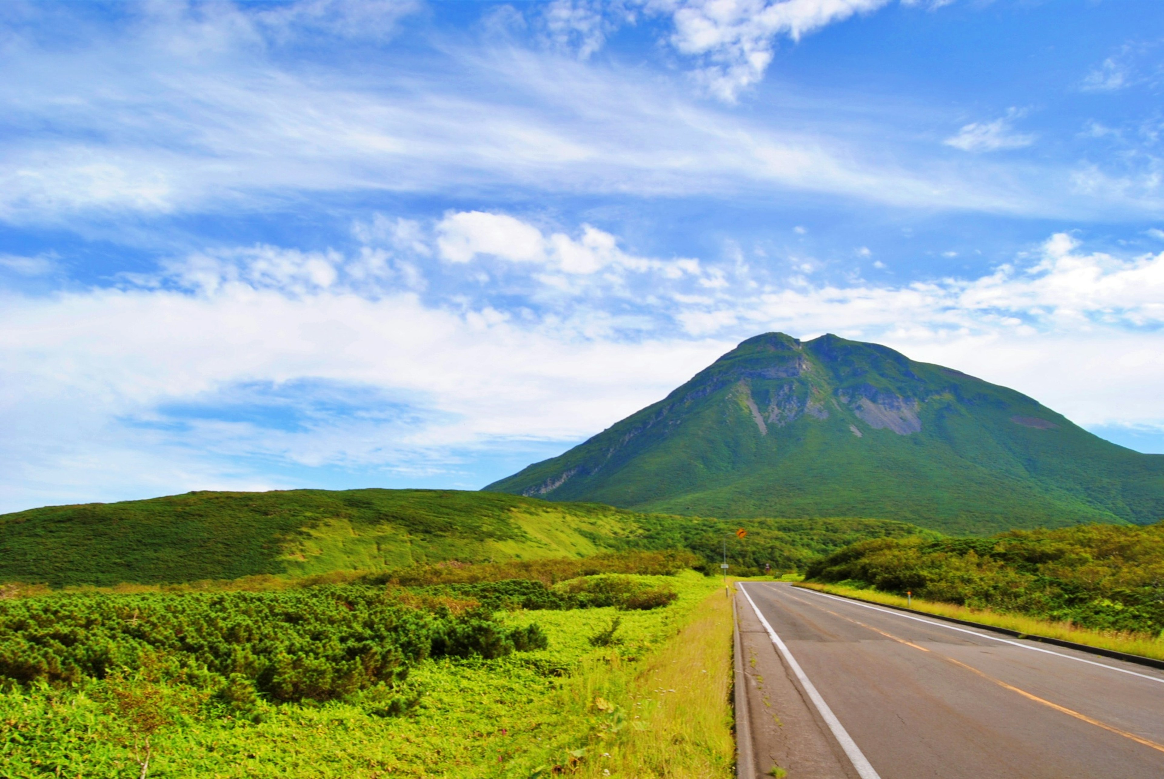 緑の草原と青い空の下にそびえる山と道路の風景