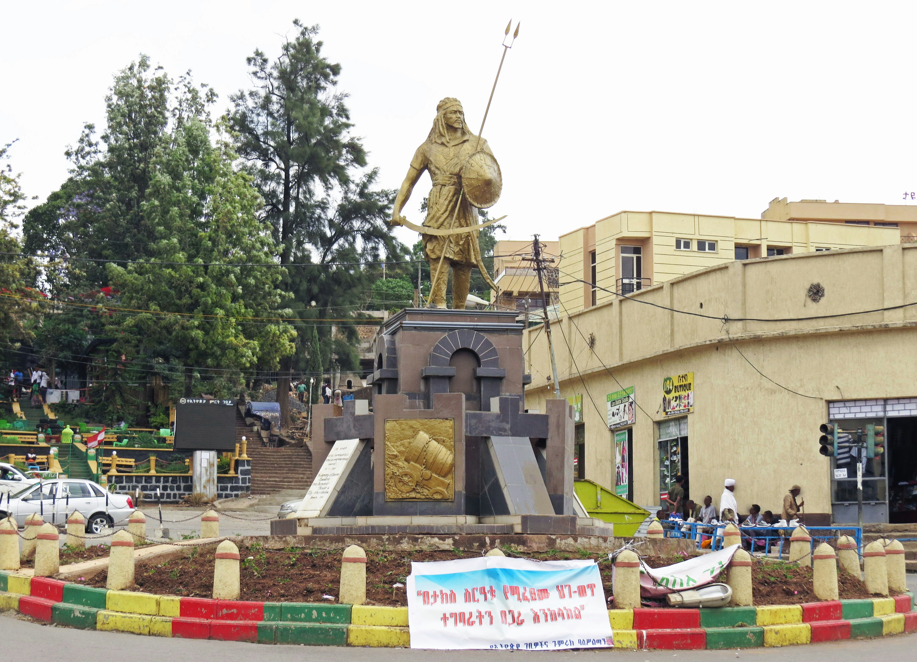 A golden statue stands in a circular plaza surrounded by green trees and buildings