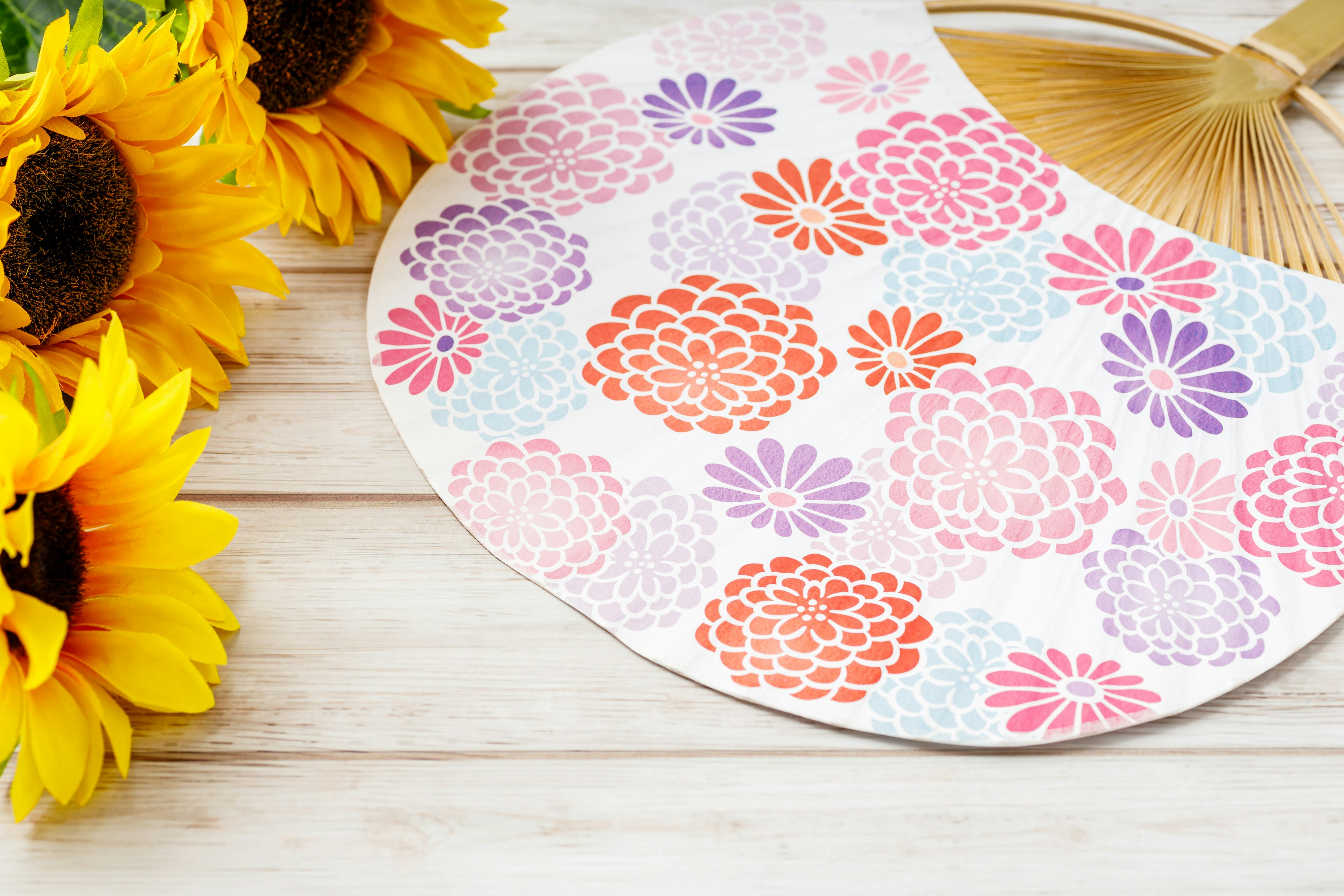 A colorful floral fan beside bright sunflowers on a wooden surface