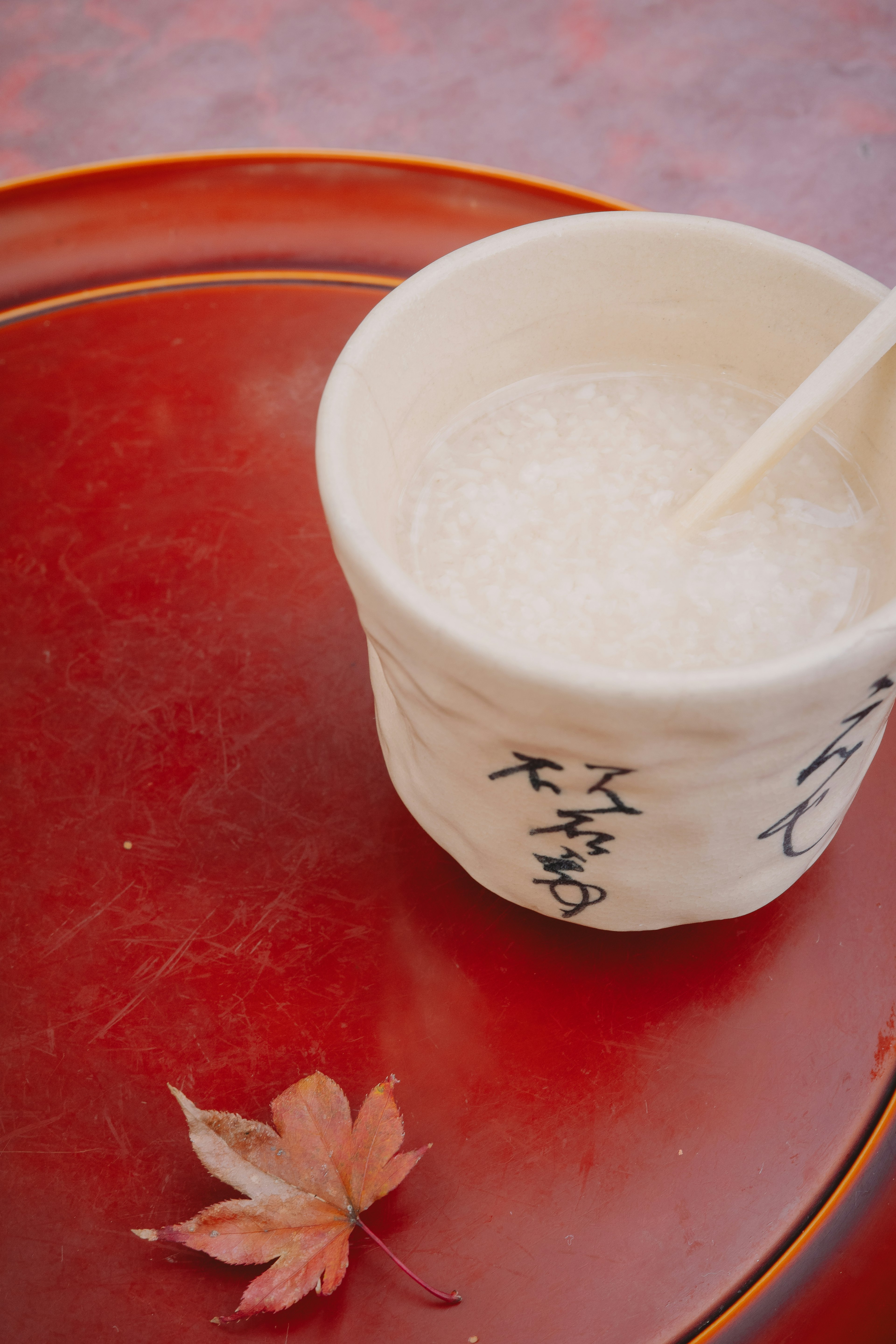 Eine weiße Tasse mit einem Reisgetränk auf einem roten Tablett mit einem roten Blatt daneben