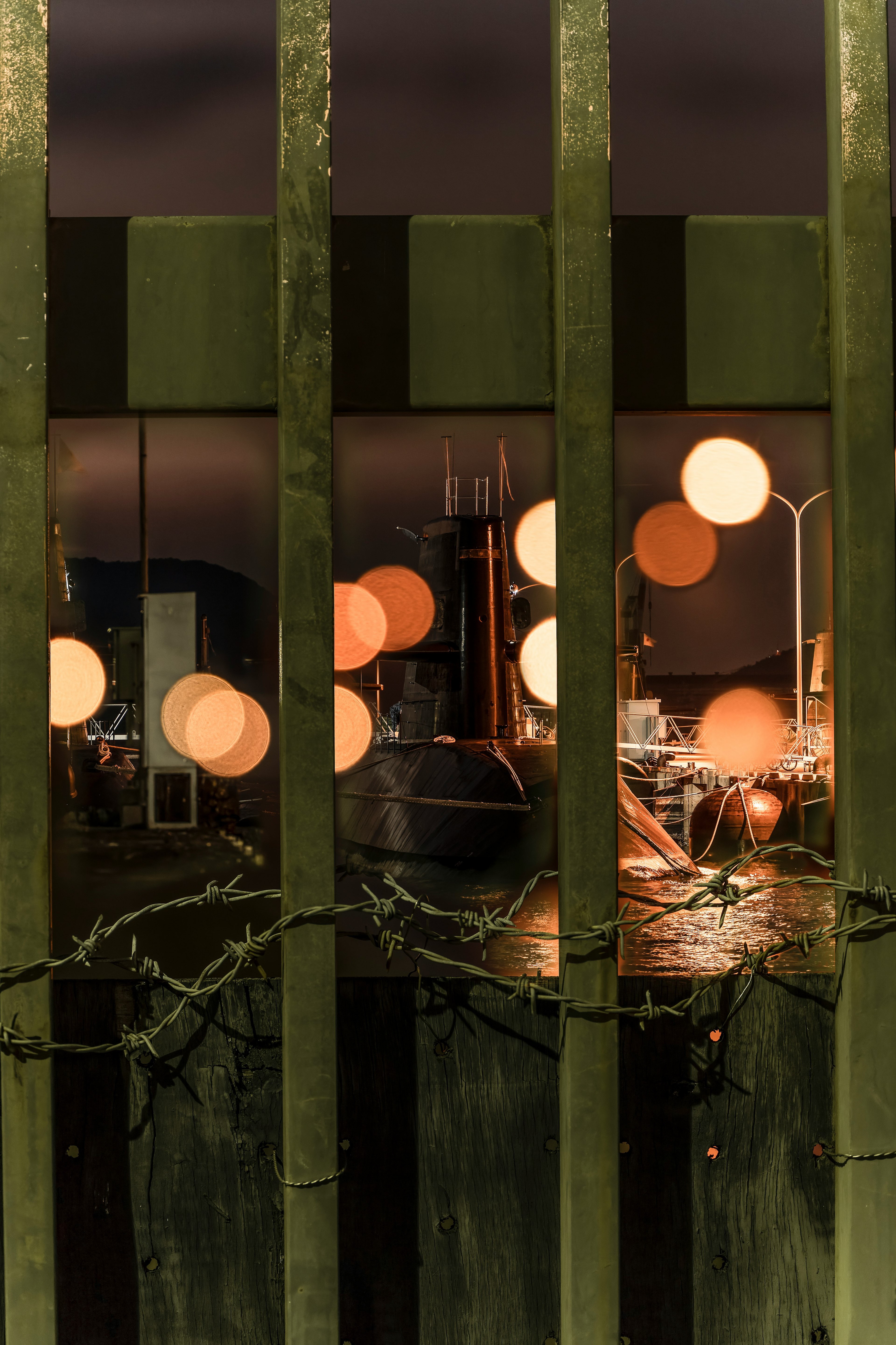 View through a green fence showcasing orange lamps and a night scene