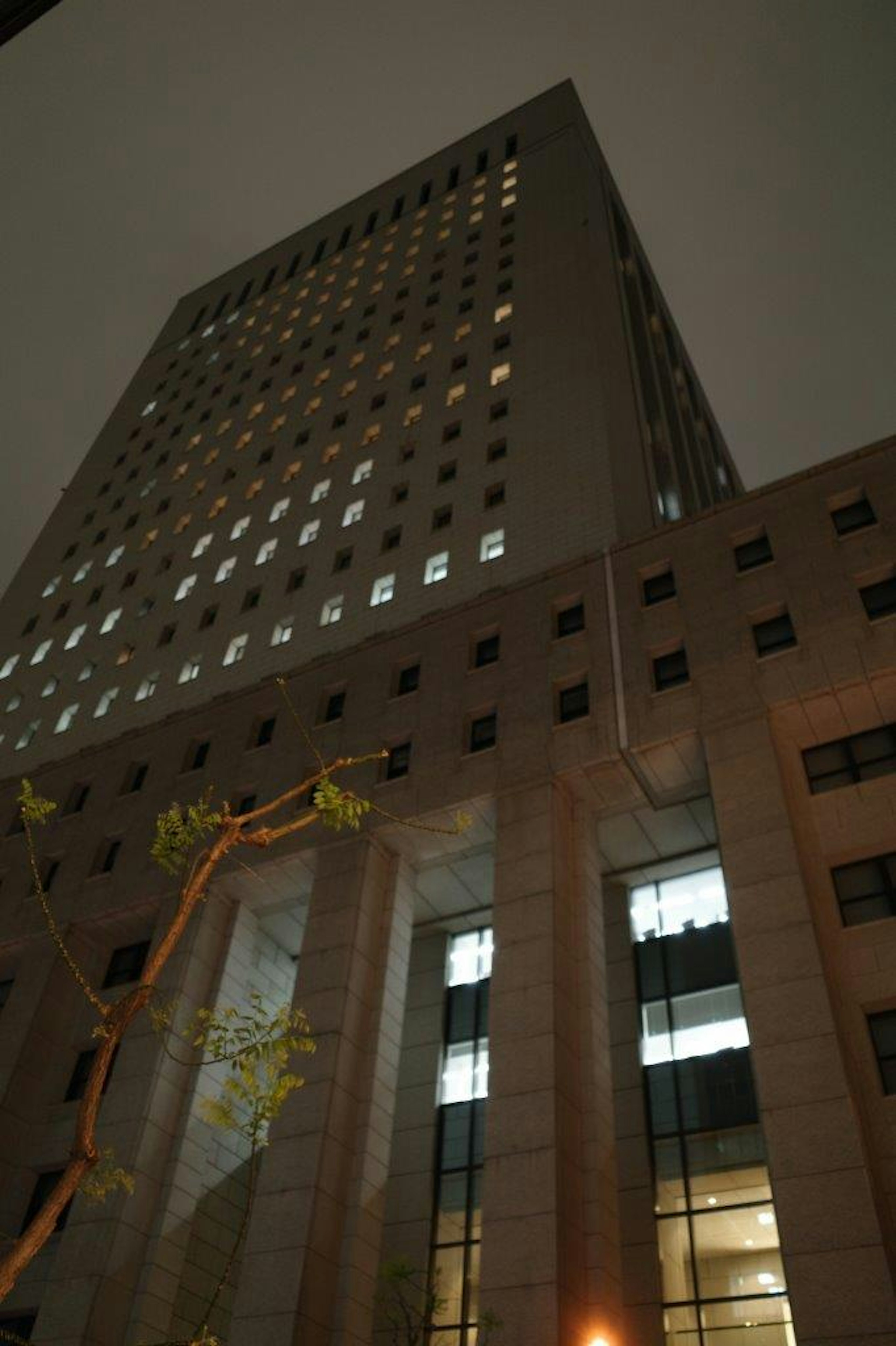 Gambar gedung pencakar langit yang diambil dari bawah pada malam hari Jendela-jendela cerah menerangi lantai atas