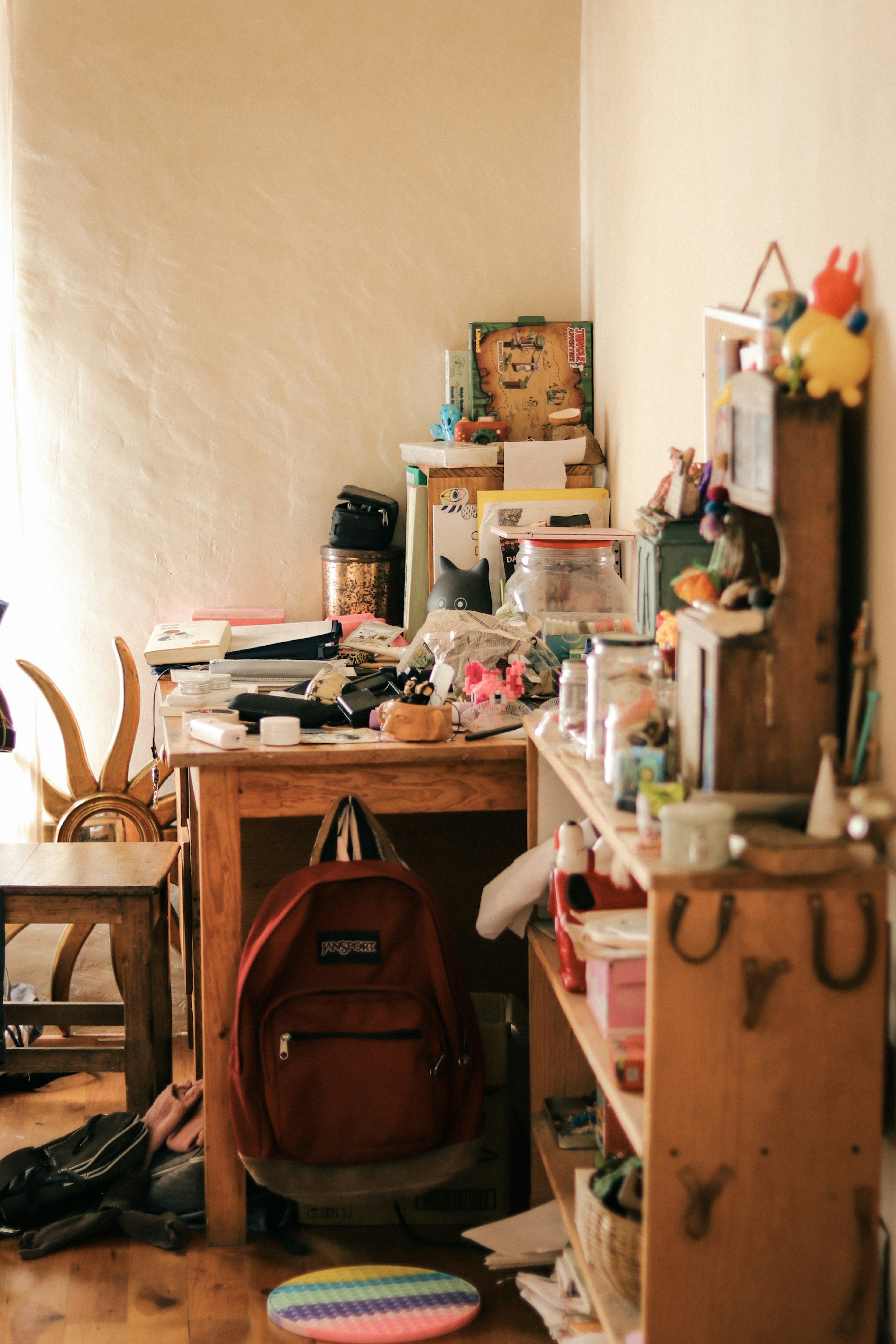 Un coin encombré d'une pièce avec un bureau en désordre et des étagères contenant un sac à dos rouge et divers objets