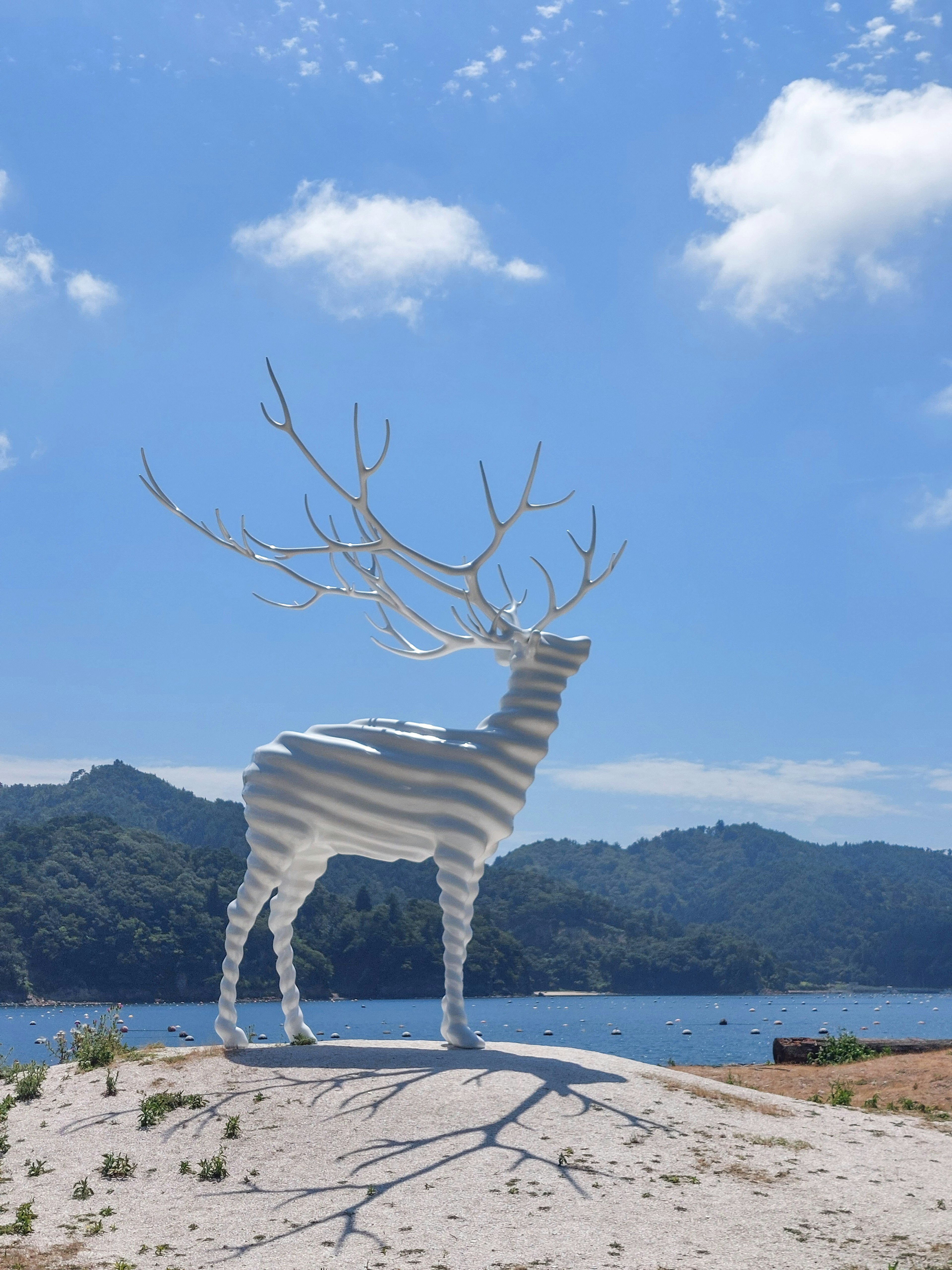 青空の下に立つ白いシカの彫刻周囲の山々と湖が見える