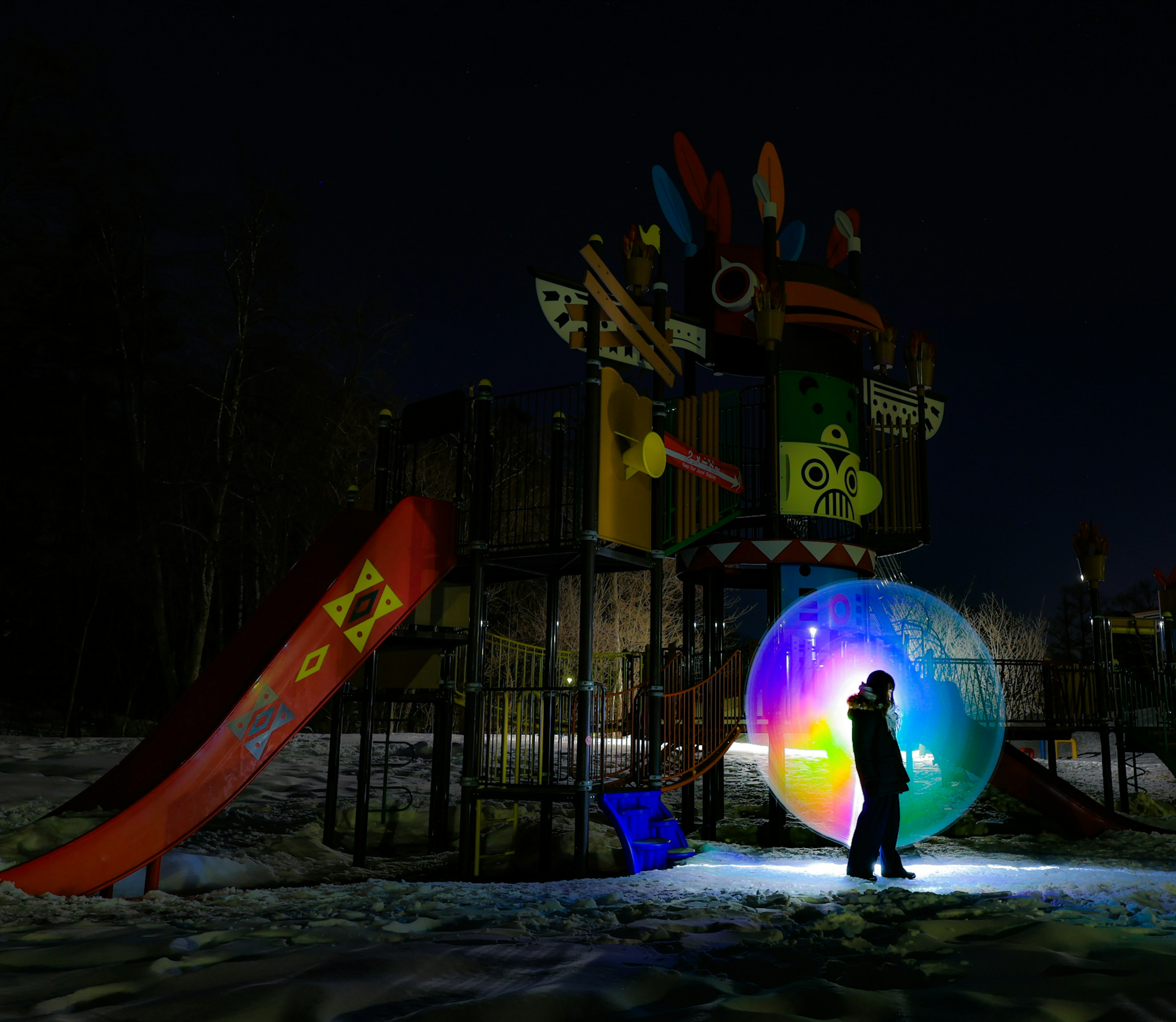 夜の公園の遊具とカラフルな滑り台のそばで光る大きな透明なボールの中に立つ人