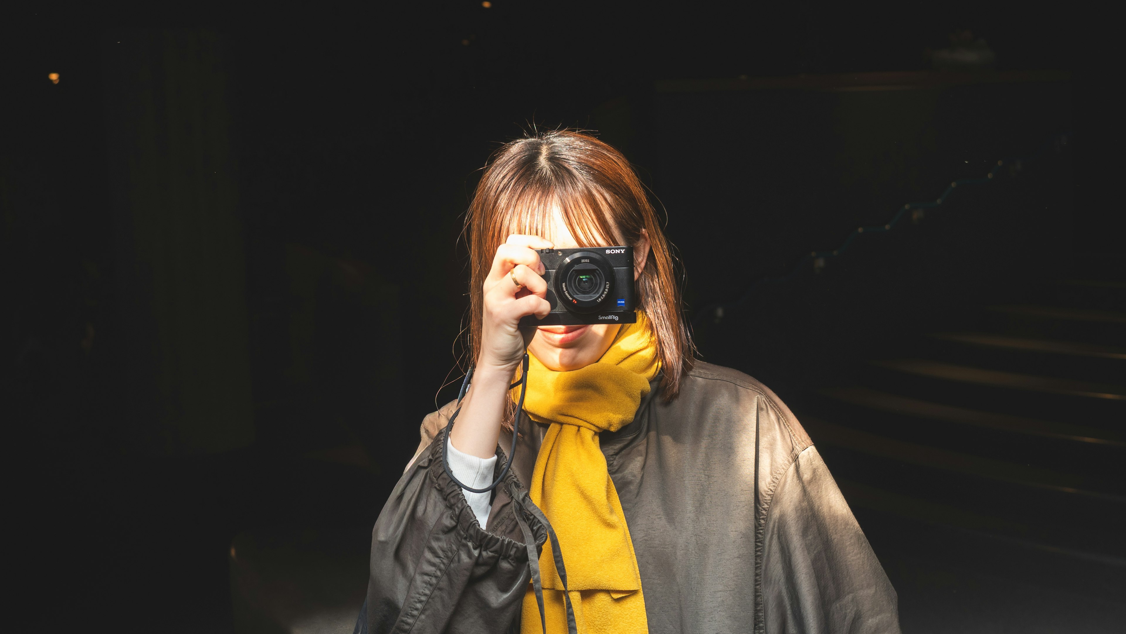 Retrato de una mujer sosteniendo una cámara contra un fondo oscuro con una bufanda amarilla