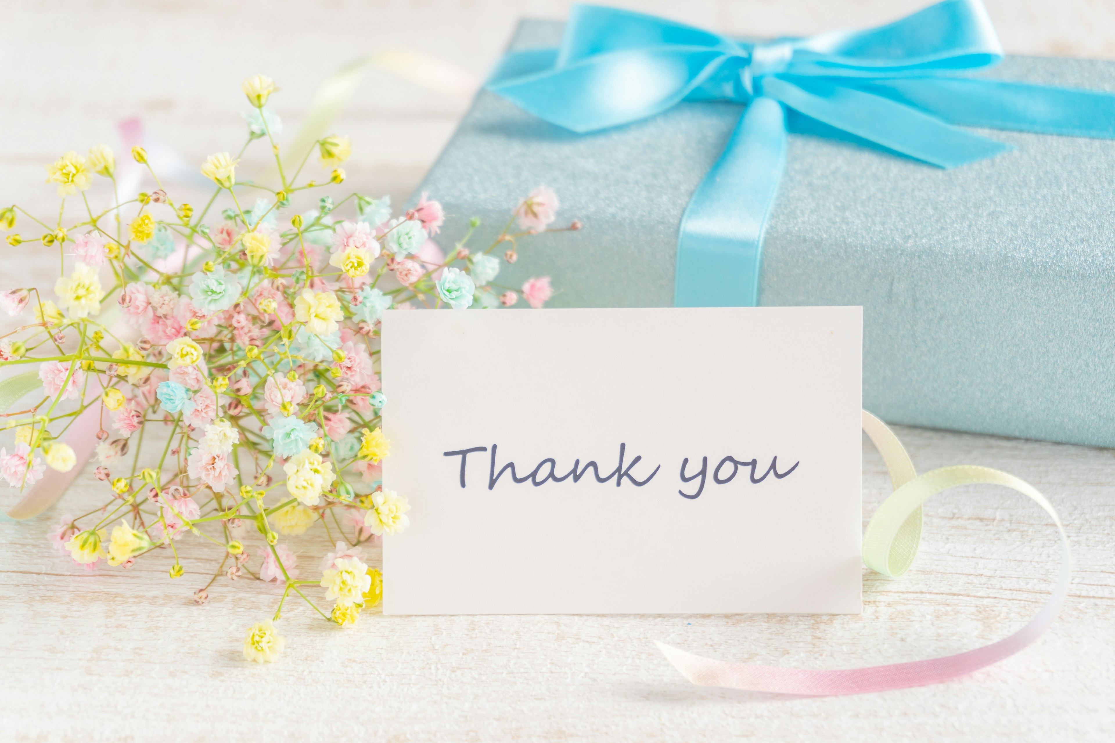 A light blue gift box with a blue ribbon and a thank you card alongside a small bouquet of pastel flowers