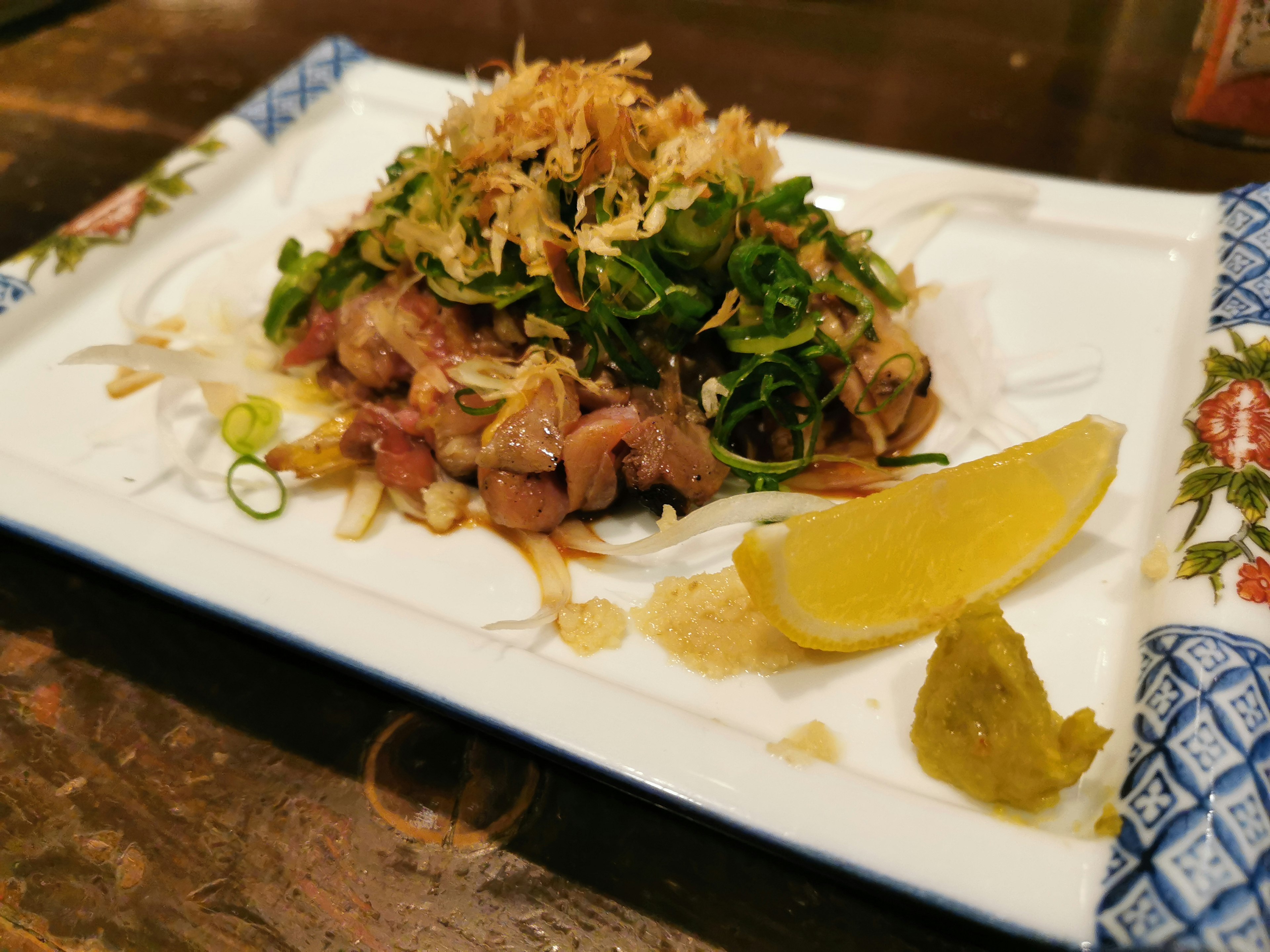 Ein Teller mit einem Gericht aus Schweinefleisch, belegt mit Kräutern und knusprigen Zwiebeln, begleitet von Zitrone und Wasabi