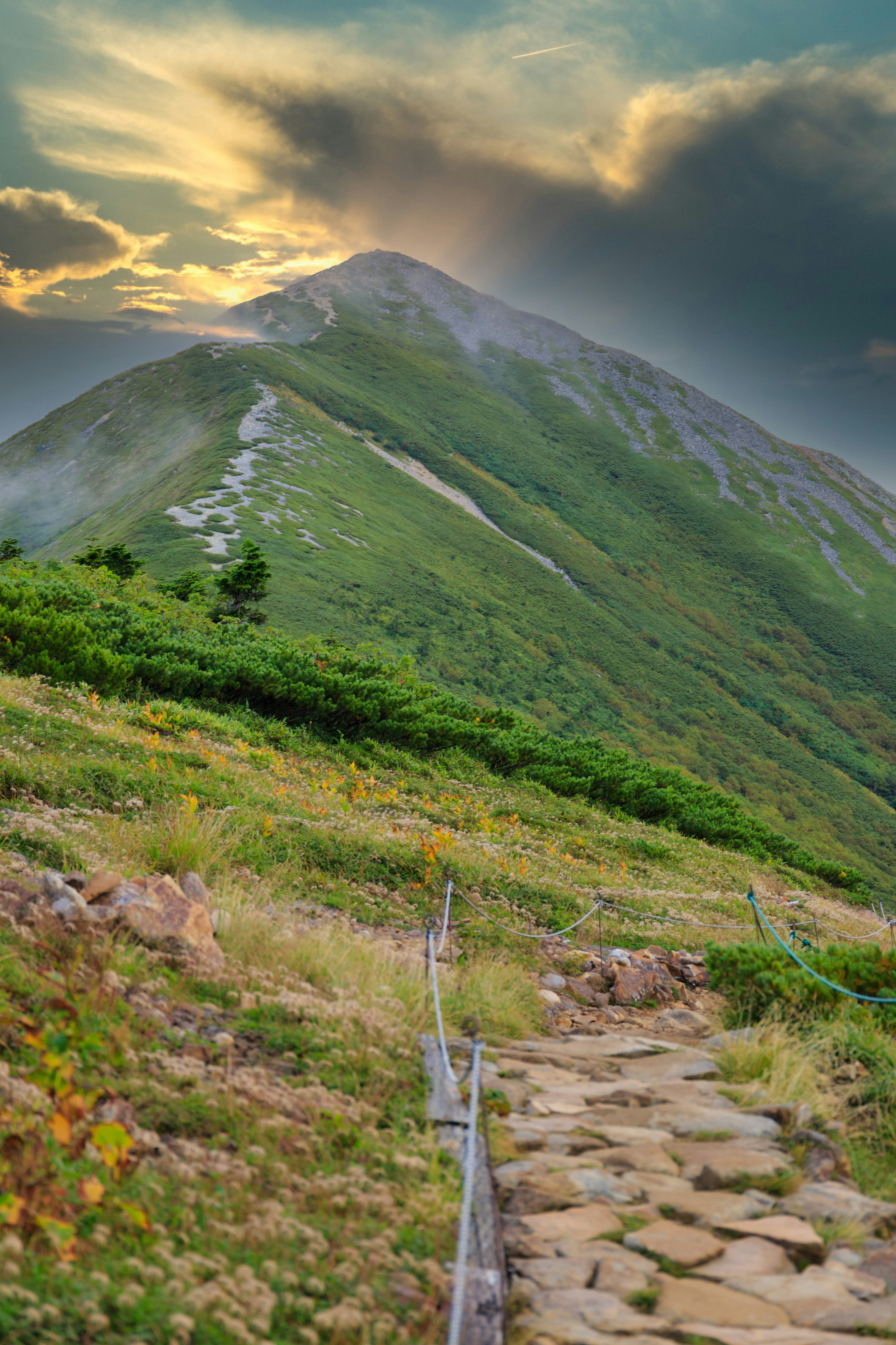 Paysage de montagne avec un chemin sinueux en pierre et une végétation luxuriante sous un ciel de coucher de soleil dramatique