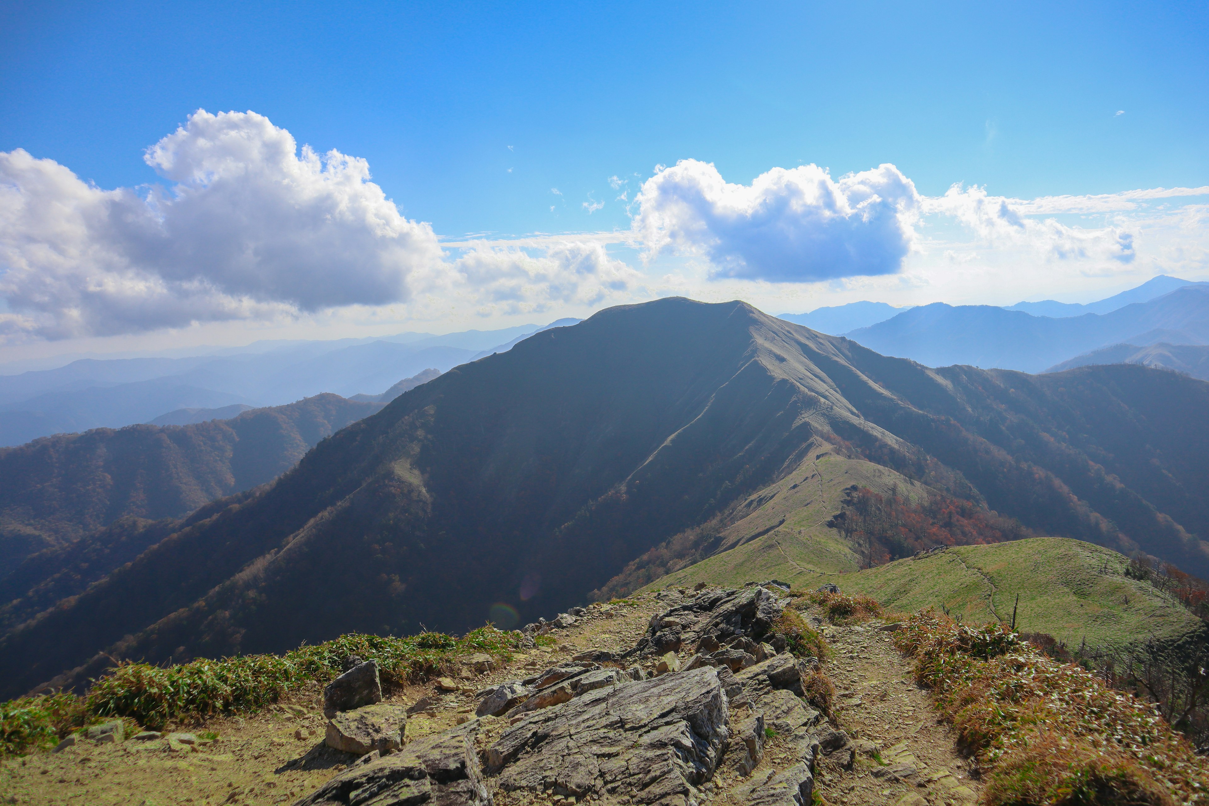 Vista desde la cima de una montaña con cielo azul, nubes blancas, colinas verdes, camino rocoso