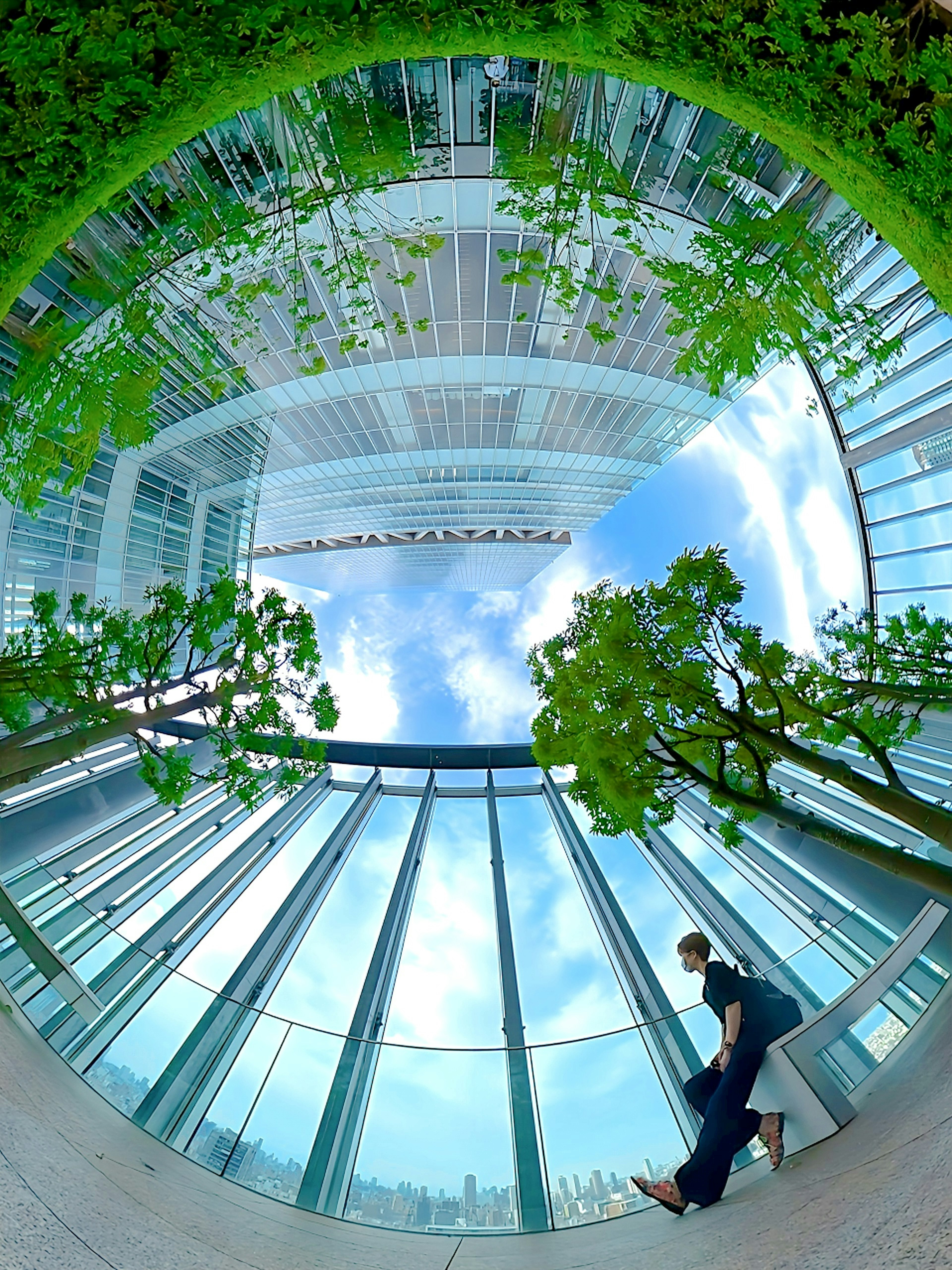 Orang yang berjalan di dalam gedung modern dengan dinding kaca yang memantulkan pohon dan langit biru