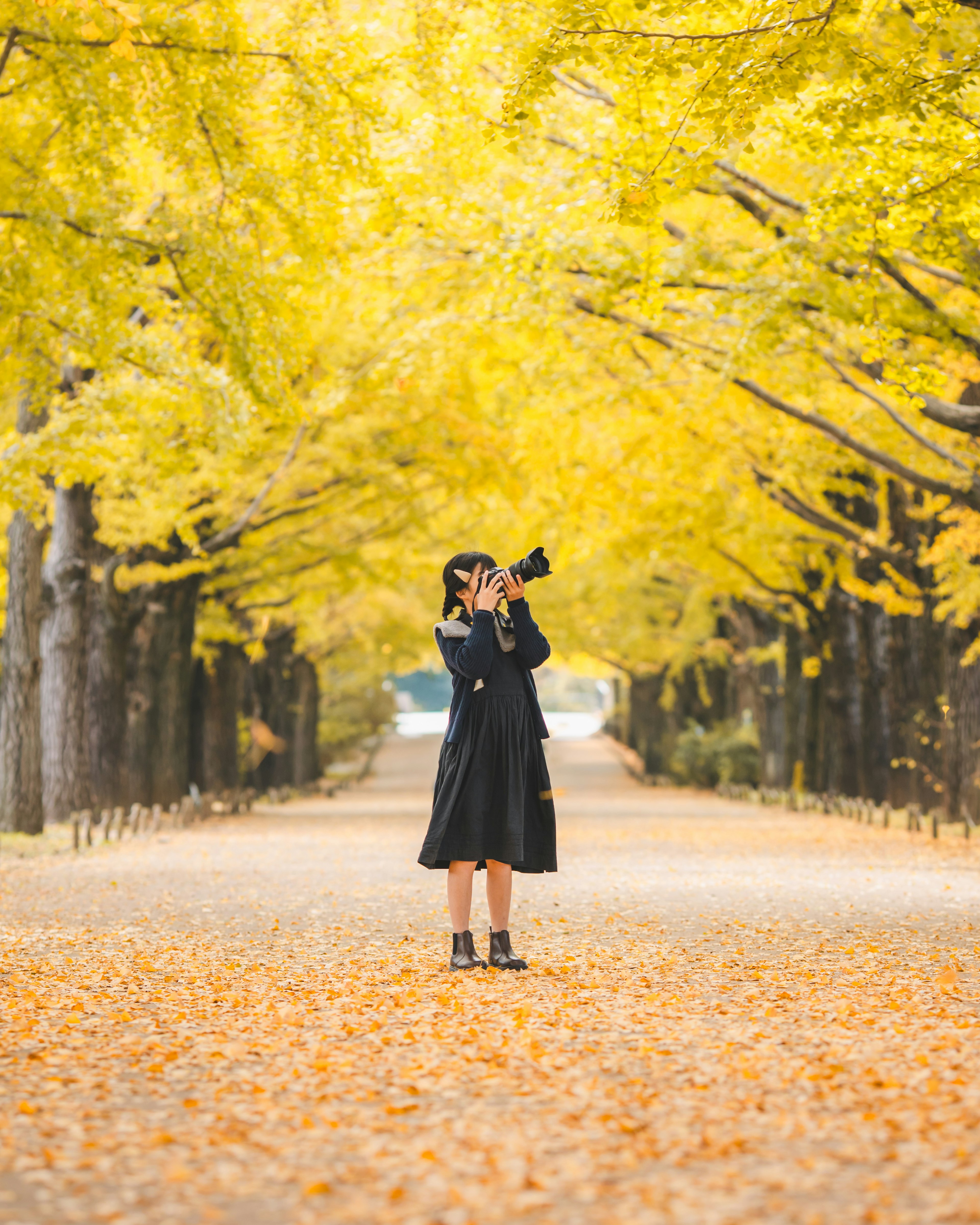 A woman standing on a path lined with yellow autumn leaves holding a camera