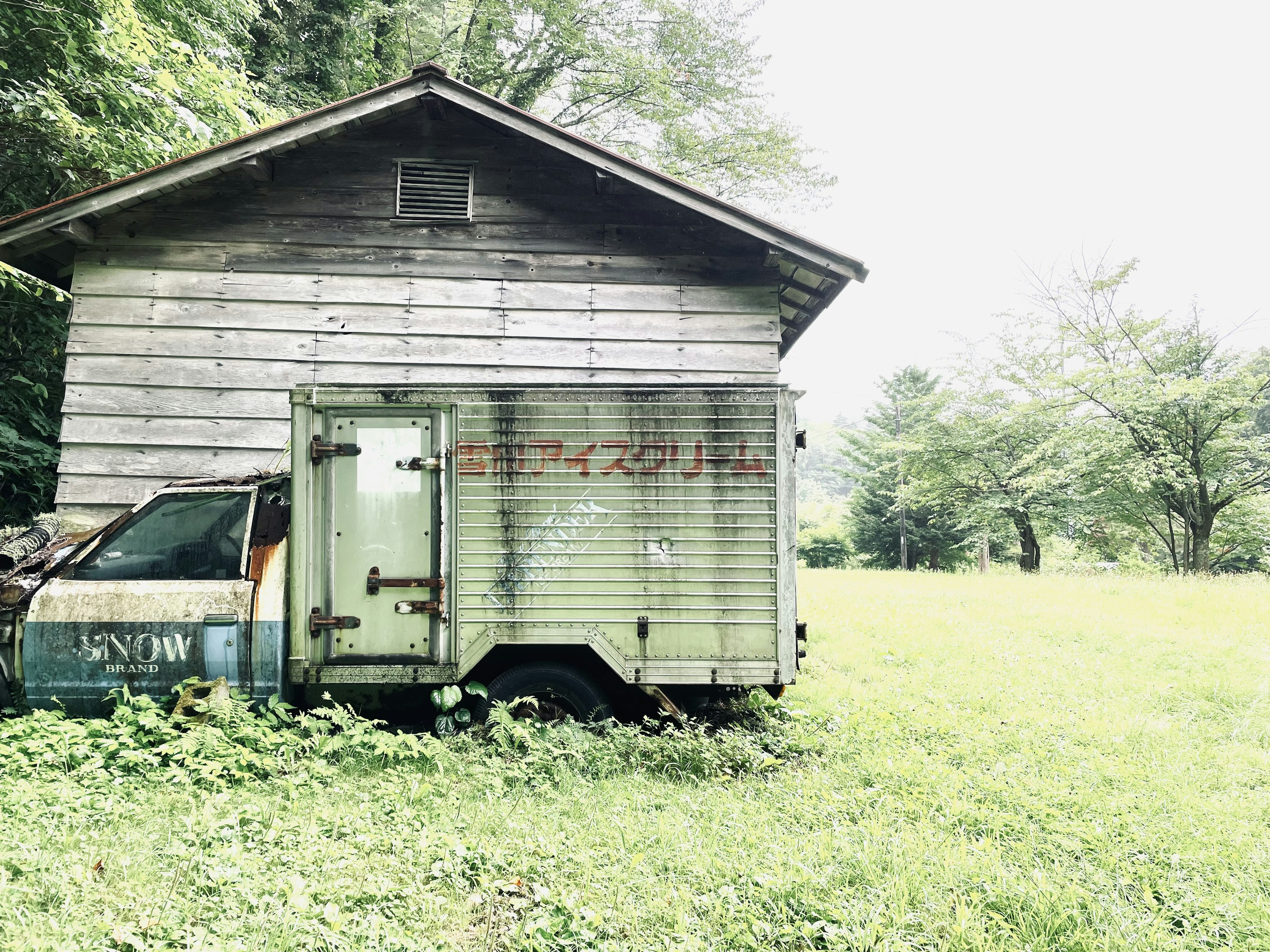 Ancienne maison en bois à côté d'une remorque verte dans un champ herbeux